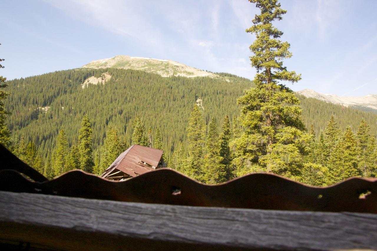 A view of a mountain landscape surrounded by dense evergreen trees, with a weathered brown roof in the foreground. The scene is set under a clear blue sky, showcasing the natural beauty of the outdoors. CDT: Alpine Tunnel mountain bike trail.