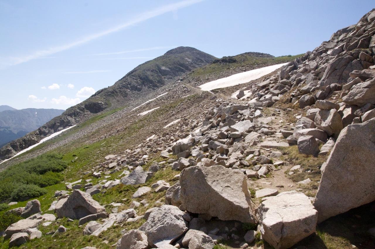 A rocky mountain trail leads upward, surrounded by boulders and patches of grass. In the background, a snow-capped peak rises under a clear blue sky dotted with a few clouds. The landscape features a mix of rocky terrain and greenery, typical of high-altitude hiking trails. CDT: Alpine Tunnel mountain bike trail.