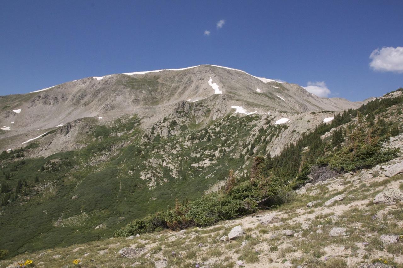 A scenic view of a mountainous landscape featuring rugged terrain, scattered rocks, and patches of green vegetation. The mountain peak is partially covered with snow beneath a clear blue sky with a few clouds. The foreground shows a grassy area with wildflowers and shrubs. CDT: Alpine Tunnel mountain bike trail.