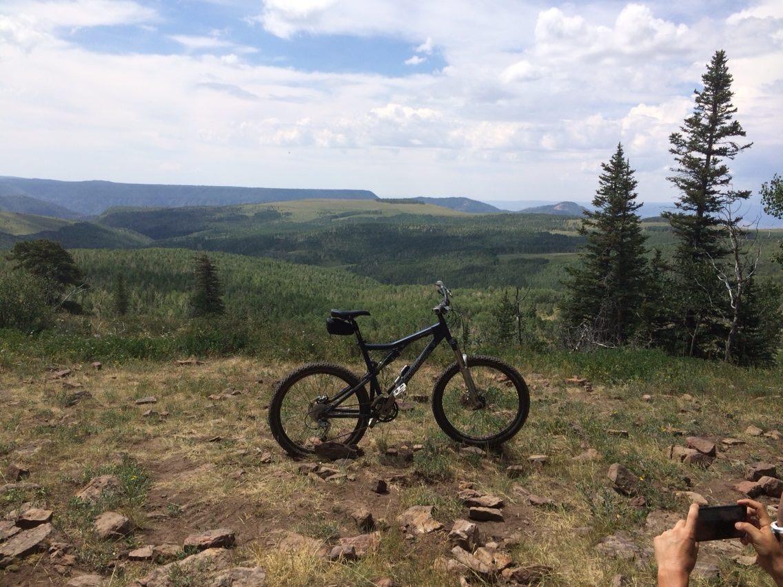 A mountain bike parked on a grassy hillside, overlooking a lush green valley and distant mountains under a partly cloudy sky. A person's hand is visible in the foreground, holding a phone, capturing the scenic view. Turkey Flats mountain bike trail.