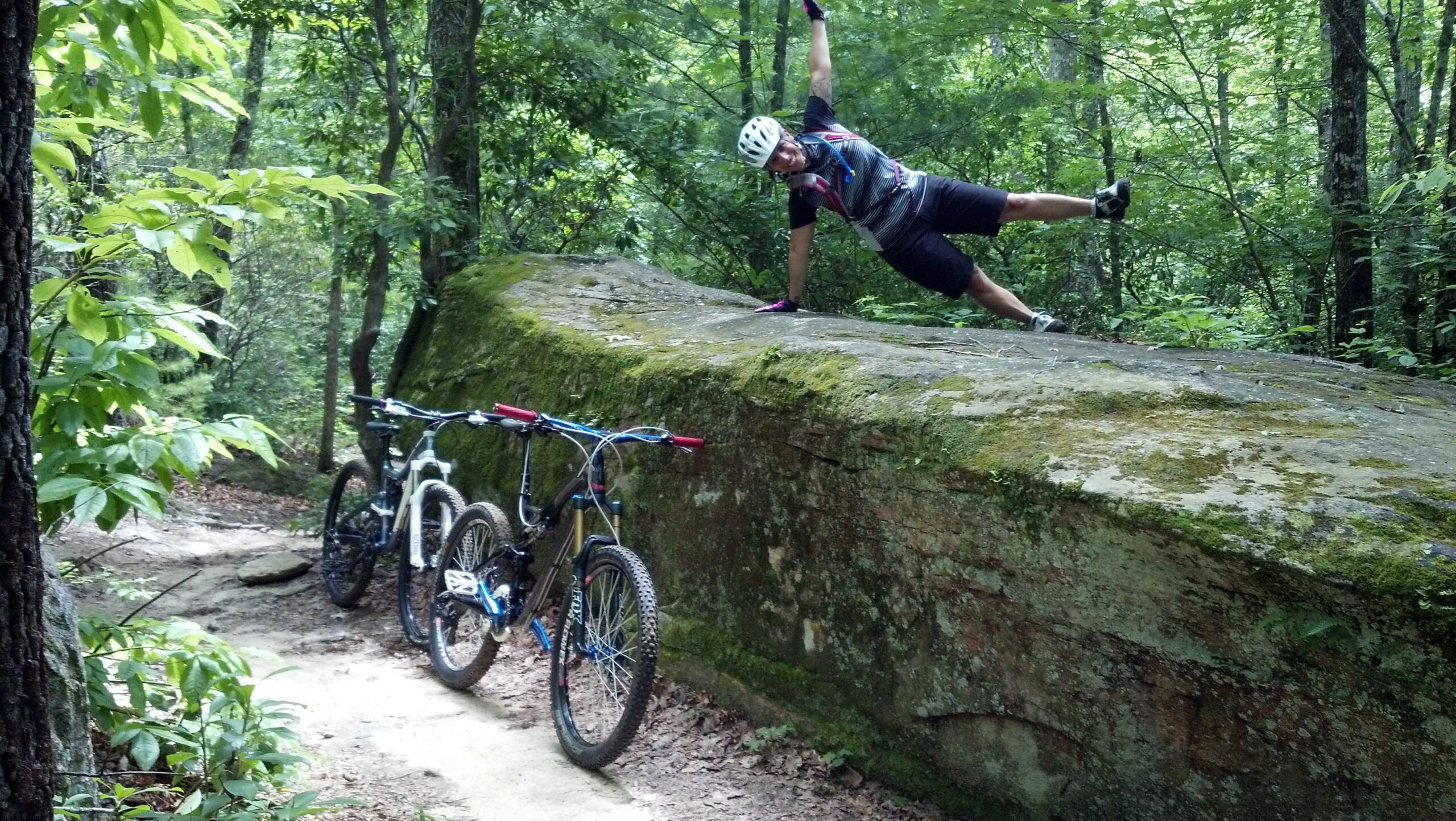 A cyclist performing a one-handed plank on a large moss-covered rock in a wooded area, with two mountain bikes parked nearby on a dirt path. The scene showcases lush greenery typical of a forest trail. DuPont State Forest mountain bike trail.