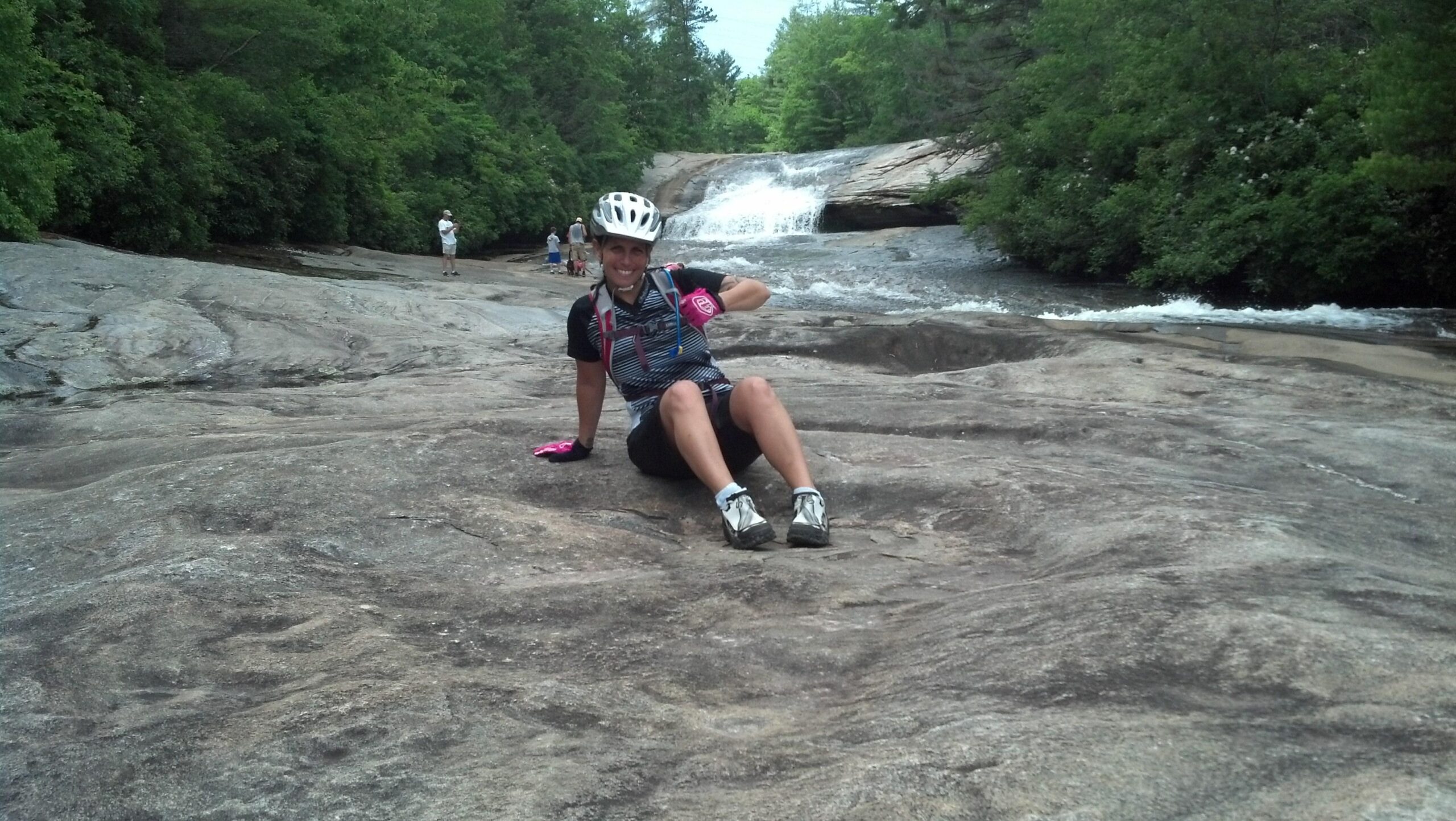 A person wearing a cycling helmet and gloves is sitting on a smooth, rocky surface next to a flowing stream. In the background, small waterfalls can be seen, surrounded by lush green trees. Other people are visible in the distance, enjoying the natural setting. The scene conveys an outdoor, adventurous atmosphere. DuPont State Recreational Forest mountain bike trail.