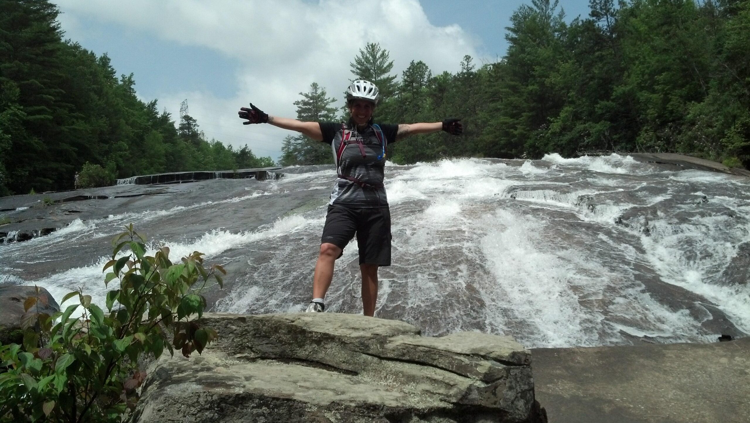 Person standing on a rock with their arms outstretched in front of a flowing waterfall, surrounded by lush greenery and a partly cloudy sky. The individual is wearing a bicycle helmet and gloves, indicating an outdoor adventure activity. DuPont State Recreational Forest mountain bike trail.
