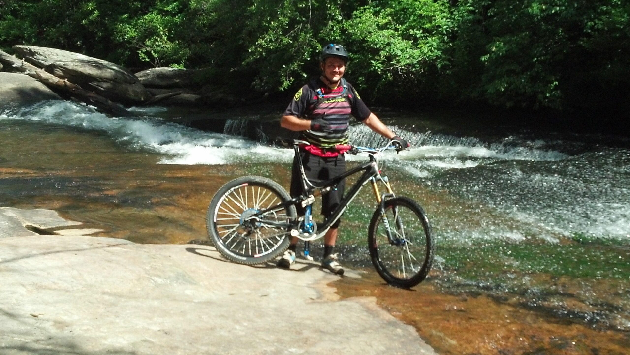 A person standing next to a mountain bike at the edge of a stream, surrounded by lush greenery and rocky terrain. The individual is wearing a helmet and casual athletic clothing, giving a thumbs up while smiling at the camera. The stream is flowing nearby, adding to the outdoor adventure vibe of the setting. DuPont State Recreational Forest mountain bike trail.