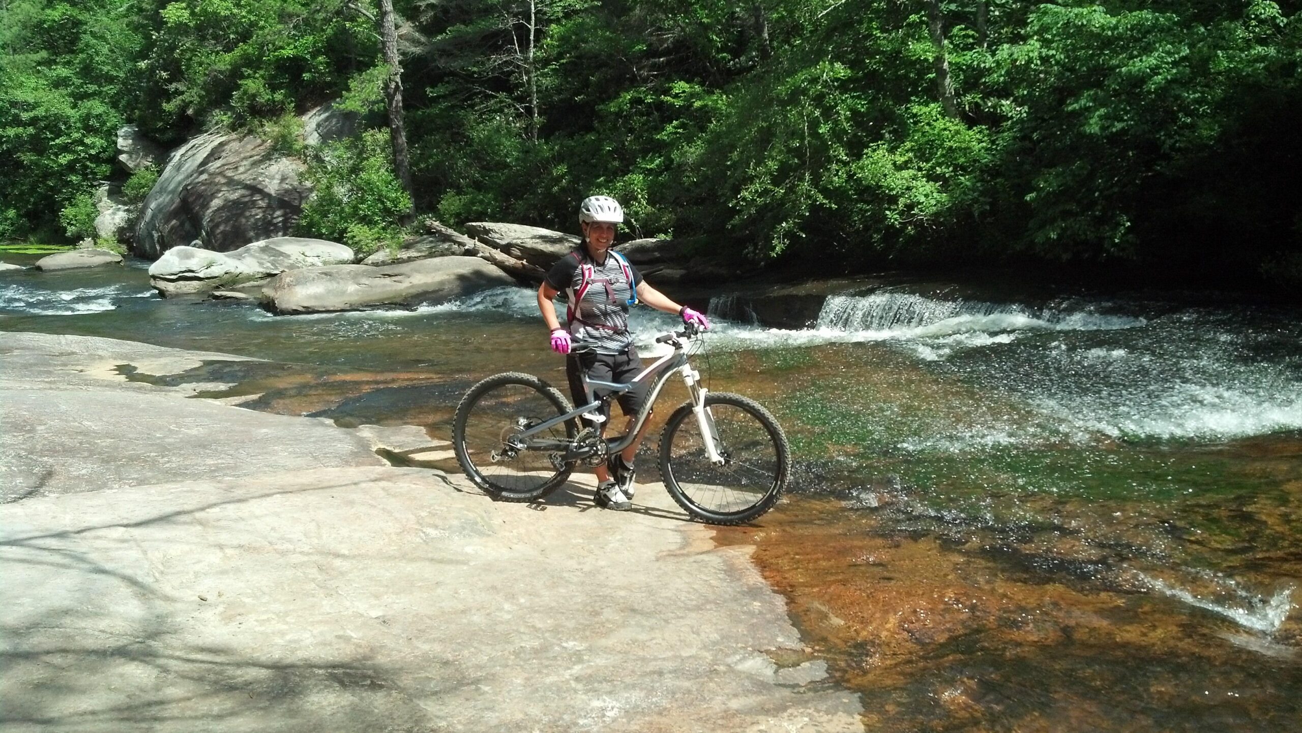 A person stands on a rocky riverbank beside a flowing stream, holding a mountain bike. They are wearing a helmet, gloves, and athletic clothing, surrounded by lush green trees and boulders. Sunlight filters through the foliage, creating a vibrant outdoor setting. DuPont State Forest mountain bike trail.