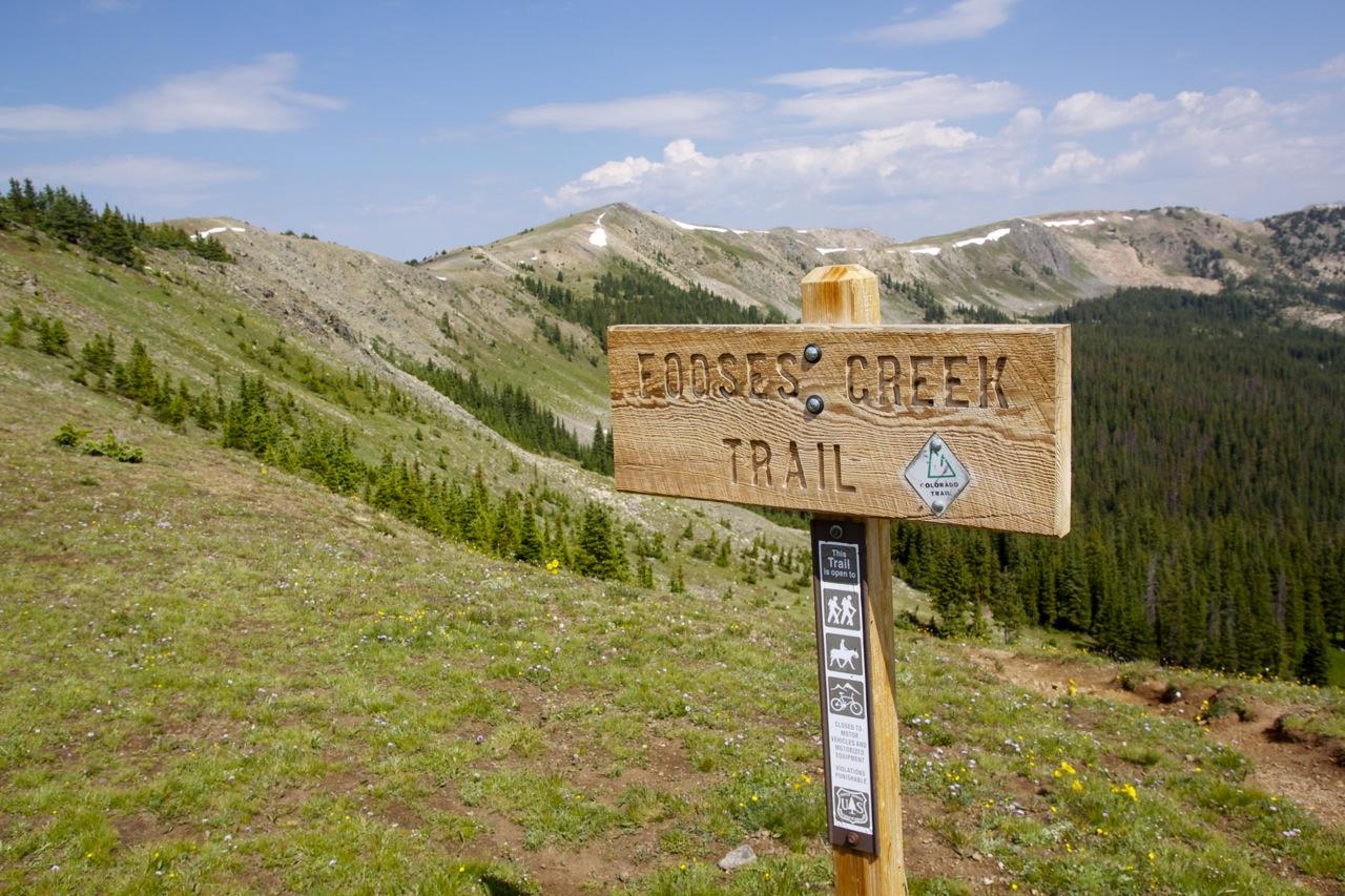A wooden trail sign reading "Fooses Creek Trail" with directional markers, set against a backdrop of rolling green hills and a partly cloudy sky. Pine trees are visible in the distance, indicating a mountainous area suitable for hiking and outdoor activities. Colorado Trail: Fooses Creek mountain bike trail.