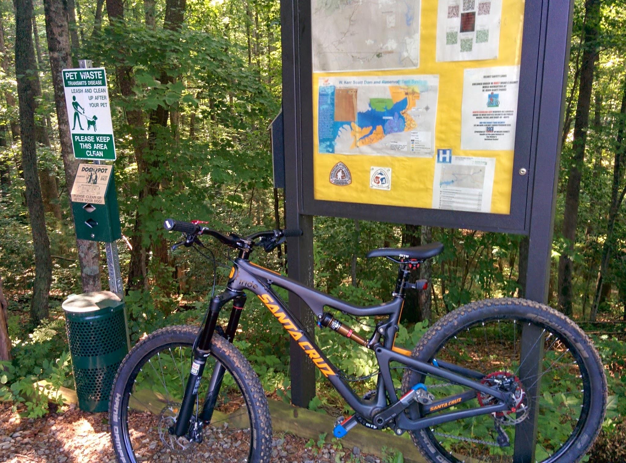 A black mountain bike parked next to a trailhead sign in a wooded area, with a "Pet Waste" sign and a dog waste station nearby. The trail map and information about the park are displayed on the sign, surrounded by lush green trees. Overmountain Victory Trail mountain bike trail.