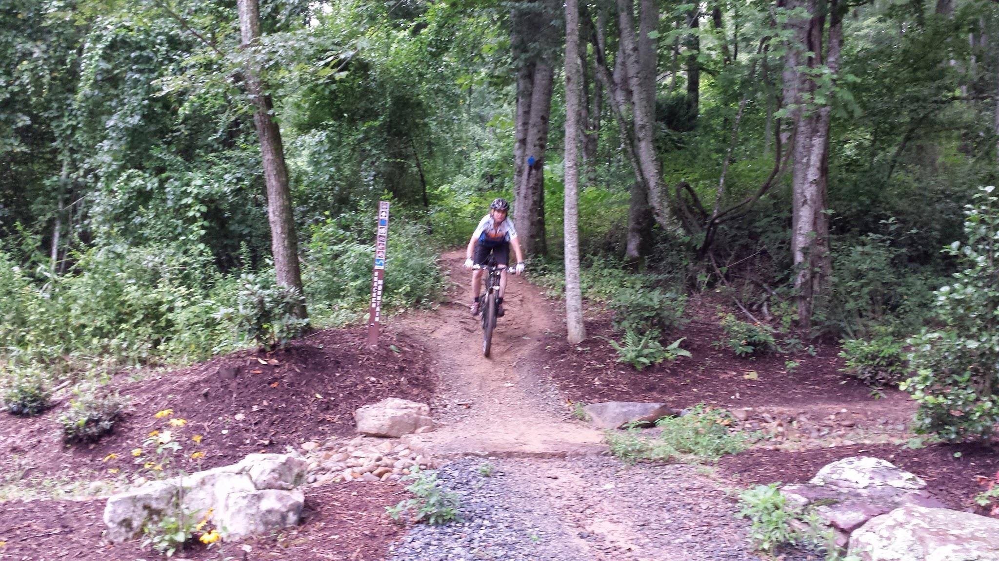 A person riding a mountain bike along a dirt path in a wooded area, navigating around trees and rocks. Lush green foliage surrounds the trail, indicating a natural setting. A trail marker is visible in the background, suggesting designated mountain biking routes. Bracken Preserve mountain bike trail.