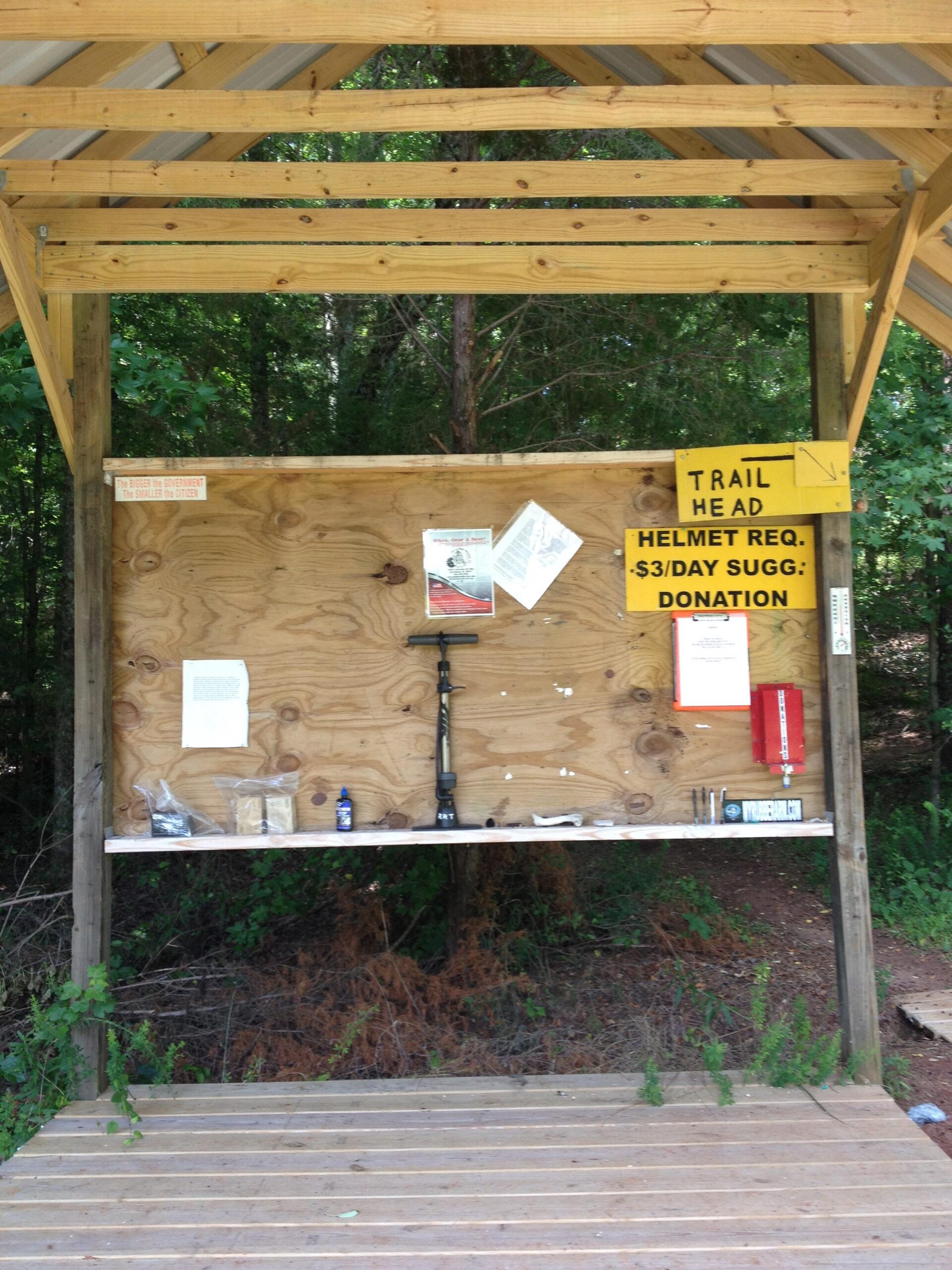 Wooden structure at a trailhead featuring a noticeboard. The board displays information about required helmets, a suggested donation, and various papers and materials related to the trail. A small shelf holds items like bottles and tools. The backdrop consists of lush greenery. Rocky River Trail mountain bike trail.