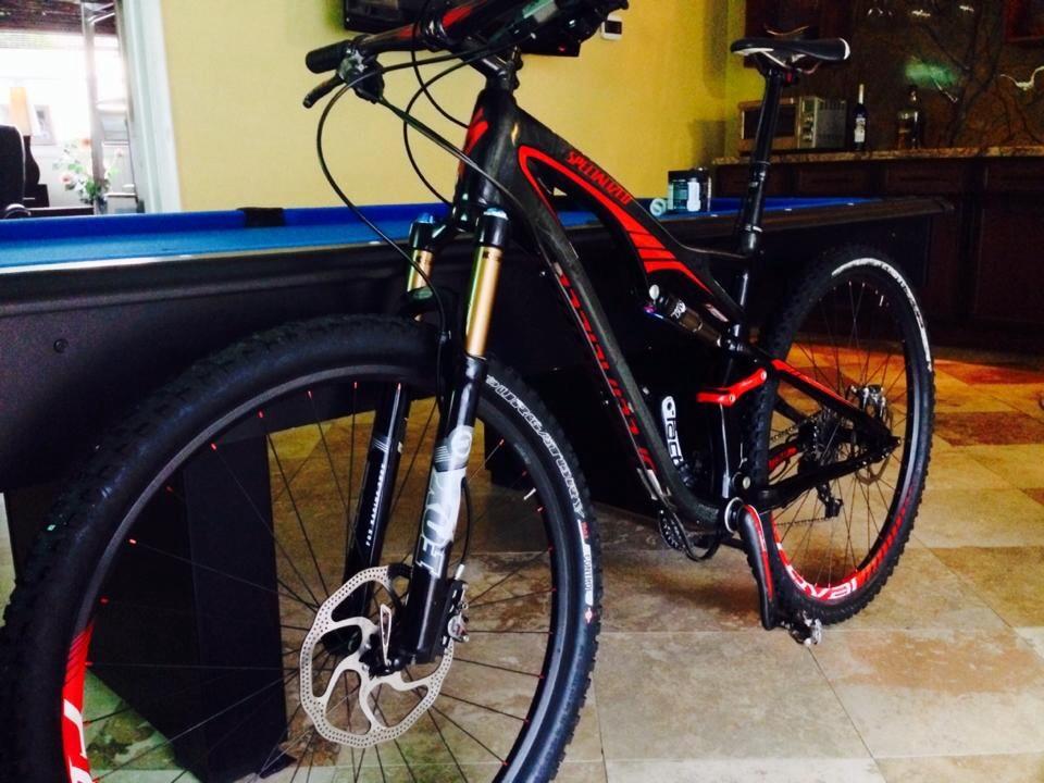 Specialized Camber Pro: A black and red mountain bike with thick tires and a front suspension fork, parked indoors next to a blue billiards table. The background features a kitchen area with cabinets and a bottle on the counter. The bike displays intricate details, including a disc brake and a gear system.