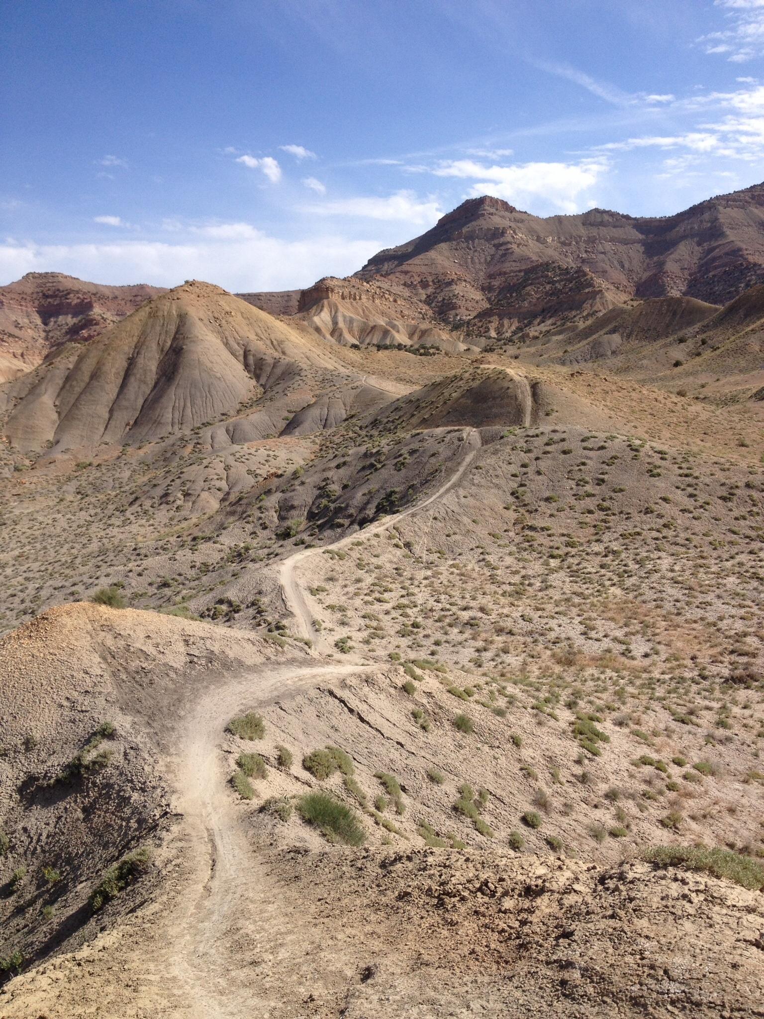 A winding dirt path leads through arid, rugged terrain, with layered hills and mountains in the background under a blue sky with scattered clouds. The landscape features sparse vegetation and a mixture of earthy tones, showcasing the natural beauty of a desert-like environment. Zippety Do Dah mountain bike trail.