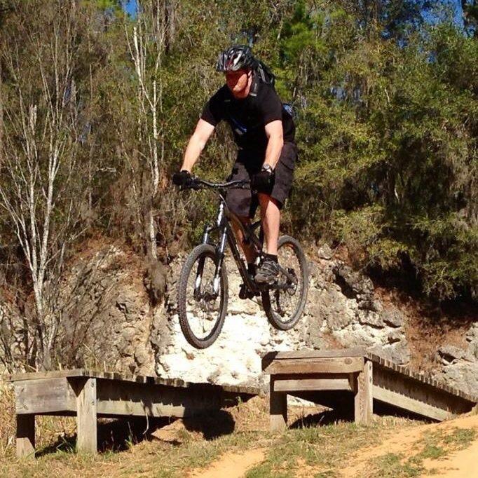 A person wearing a helmet and black clothing is jumping with a mountain bike over two wooden ramps in a natural outdoor setting. Trees and a rocky backdrop are visible in the scene, indicating a mountain biking trail. Santos mountain bike trail.