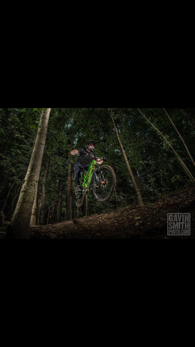 Santa Cruz Nomad Carbon: A mountain biker in mid-air, performing a jump over a dirt path in a dense forest. The rider is wearing a helmet and protective gear, and riding a bright green bike. Trees and foliage surround the area, creating a natural, adventurous setting.