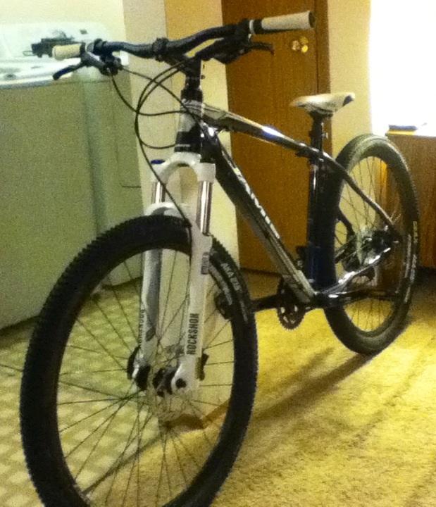 Jamis Nemesis: A mountain bike positioned indoors on a carpeted floor, featuring a dark frame and white front suspension fork. The bike has a sleek design with thick tires, handlebars with grips, and a prominent seat. In the background, there is a laundry area with a washing machine and a wooden door.