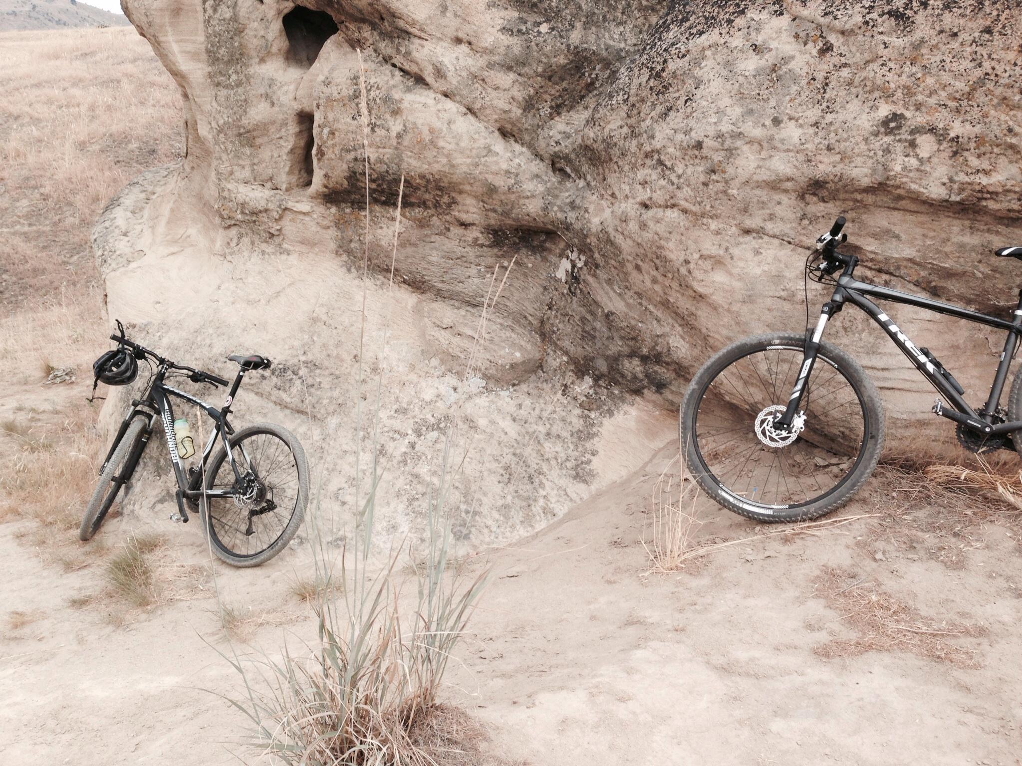 Two mountain bikes rest against a rocky outcrop in a dry, grassy landscape. The scene features muted earth tones, with the bikes positioned on a sandy path near the base of the rocks. Tall grass gently sways nearby, suggesting a serene outdoor setting ideal for biking adventures. Sage Hills mountain bike trail.