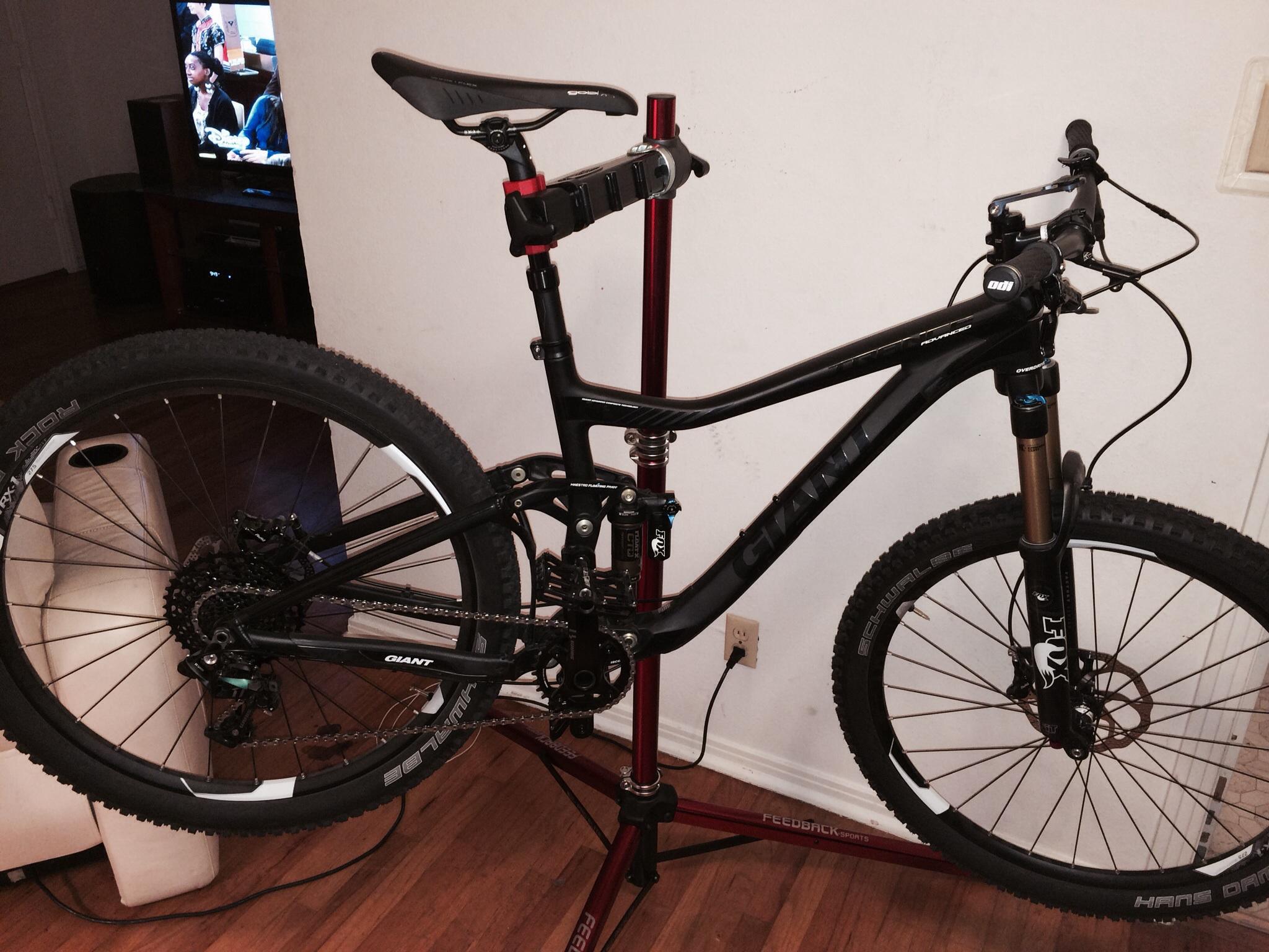 Giant Trance Advanced SX 27.5: A black mountain bike is mounted on a bike repair stand, showcasing its frame and components. The background features a television, and the flooring is wooden.