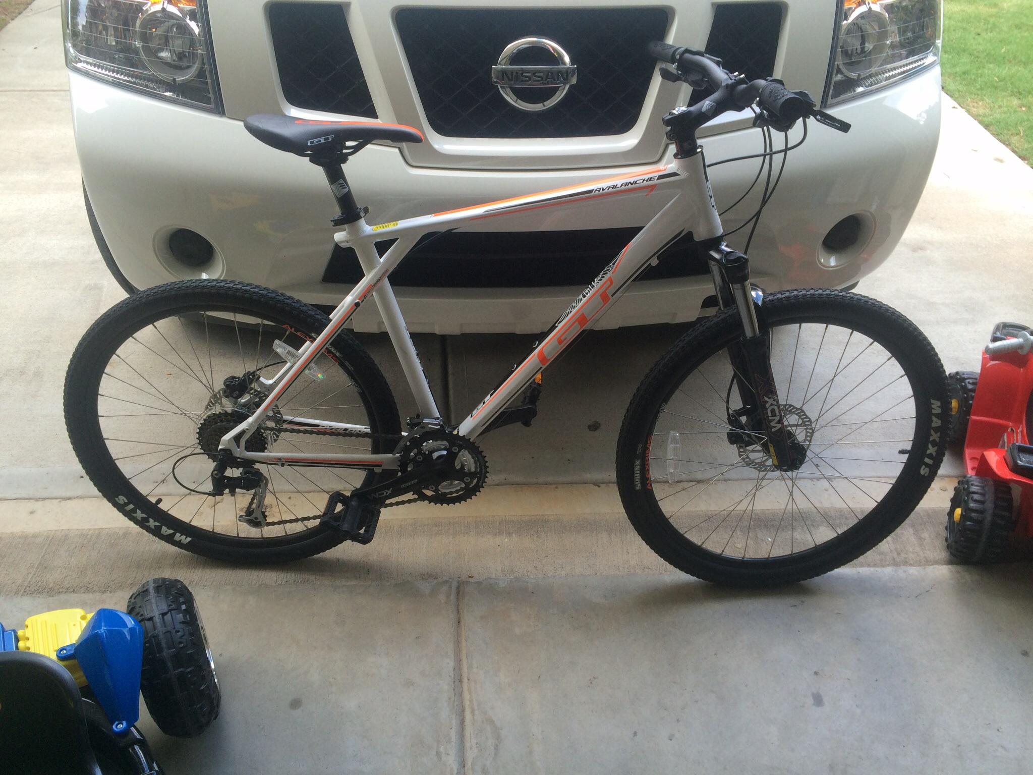 GT Avalanche: A mountain bike with a white frame and orange accents parked in a garage, with a Nissan vehicle visible in the background and toy vehicles on the side.