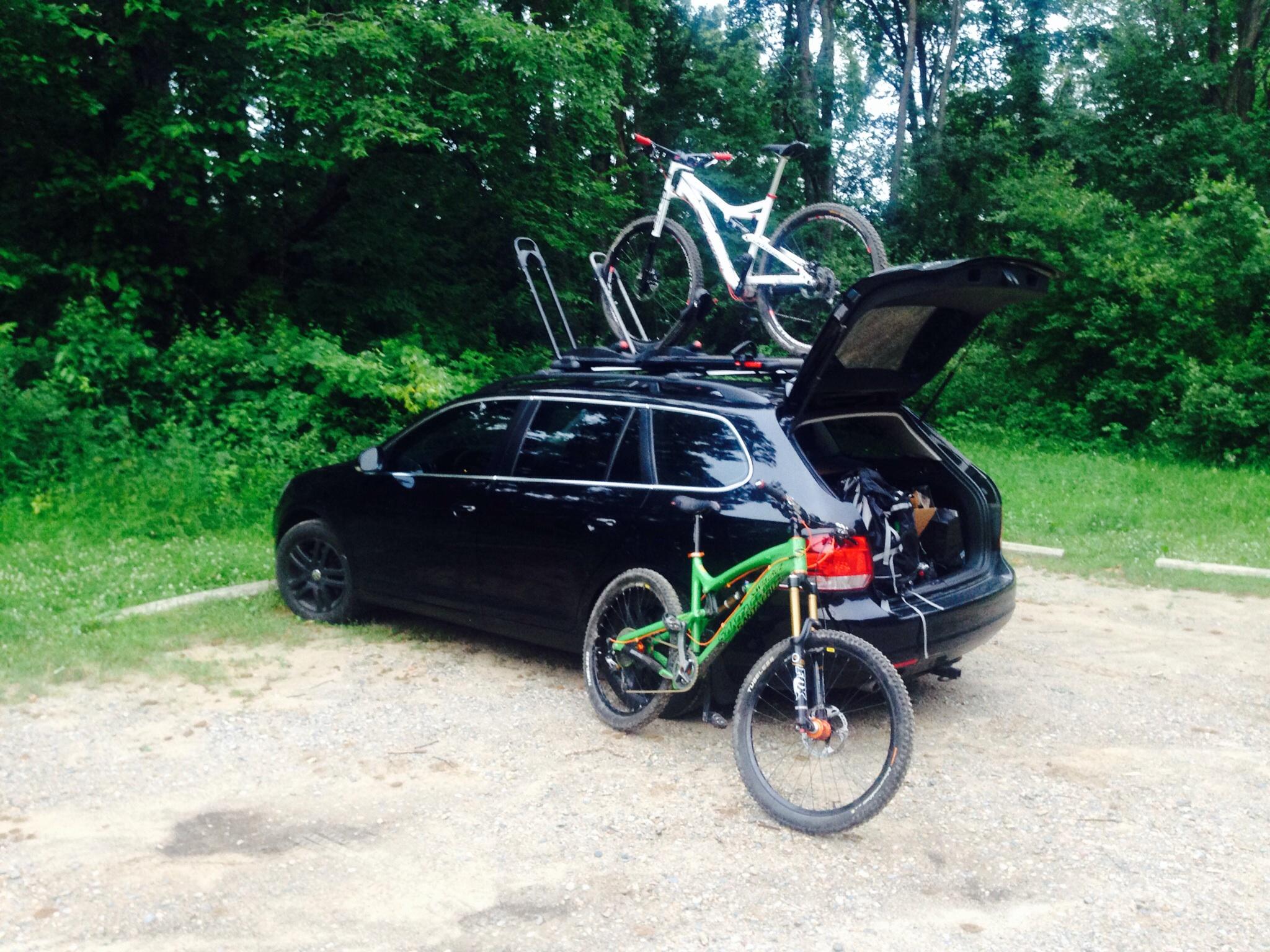 Cannondale Scalpel 29er 1: A black car parked on a gravel surface with a mountain bike mounted on a roof rack and another bike leaning against it. The car's hatchback is open, revealing bags and equipment in the trunk. Lush green trees and foliage surround the area, indicating a natural setting.