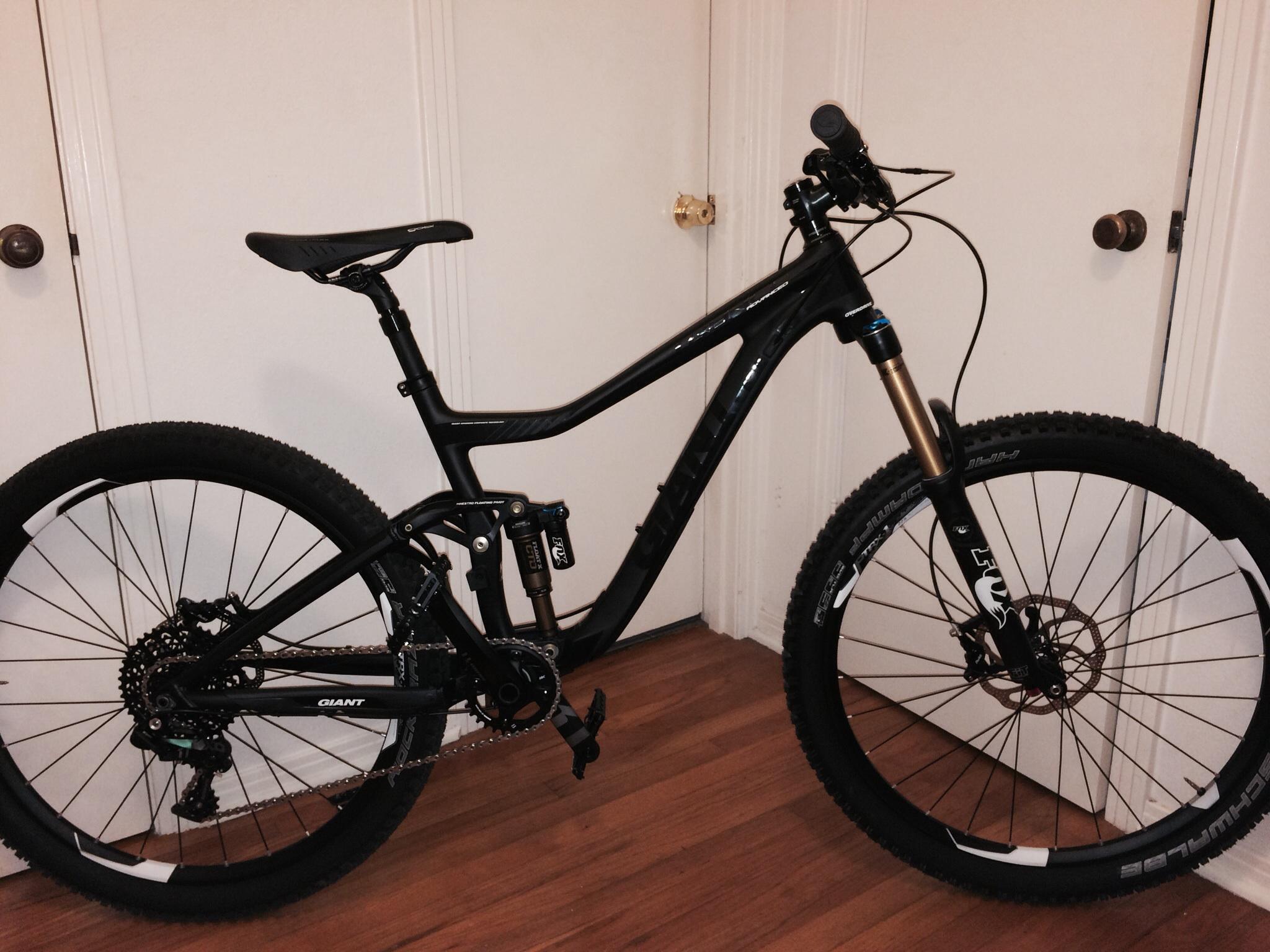 Giant Trance Advanced SX 27.5: A black mountain bike leaning against a wall with wooden flooring. It features a sleek frame, prominent suspension components, and wide tires designed for off-road riding. The handlebars are equipped with grips and a small electronic device. The background shows a closed door with a simple door knob.