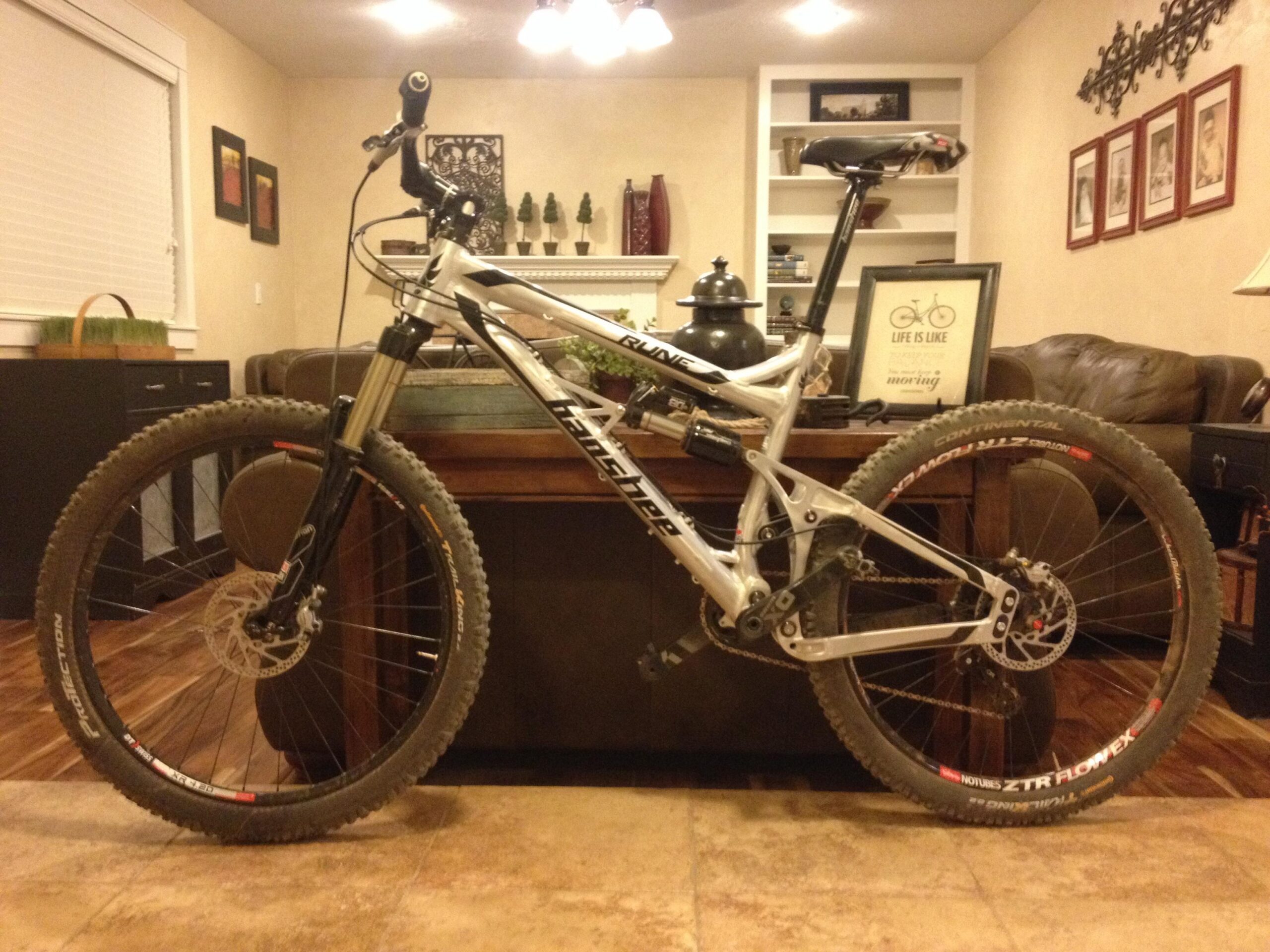 Banshee Rune: A mountain bike with a silver frame and knobby tires parked inside a living room. The bike features a front suspension fork and various components visible. Behind it, there is a coffee table with a framed motivational quote about biking, and a couch with decorative elements in the background.