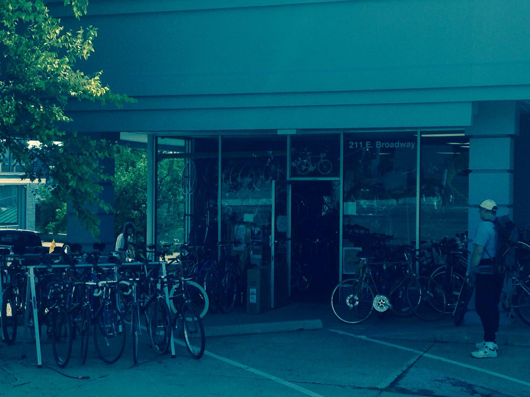 A bicycle shop exterior on a sunny day, featuring a large display of bicycles outside and a glass storefront. A person is seen standing near the entrance, while several bikes are parked in front. The address "211 E. Broadway" is visible on the building. Green trees are in the background, suggesting a vibrant, outdoor atmosphere.