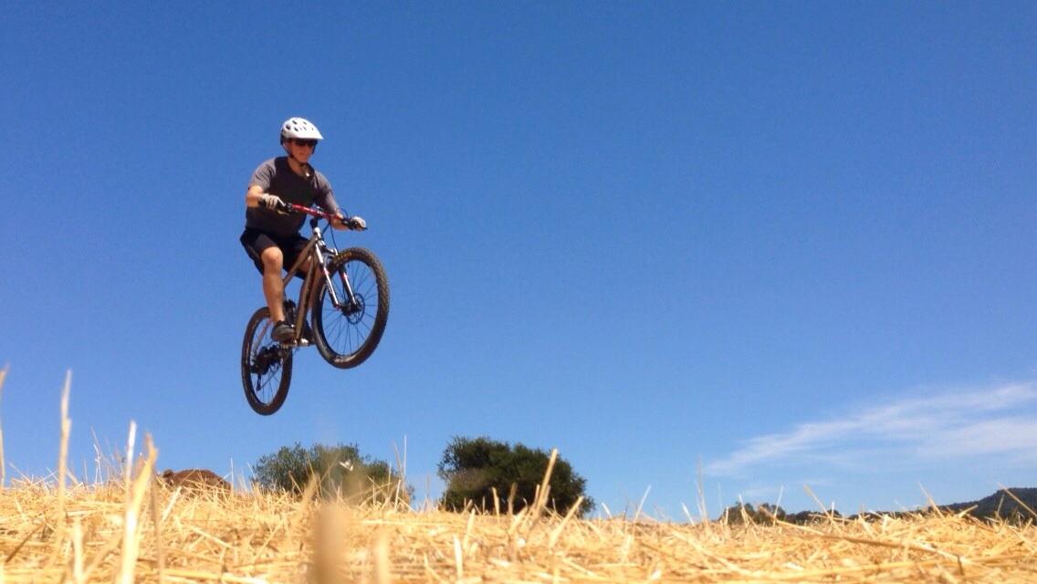 Marin Rift Zone Xc8 2013: A person wearing a helmet is performing a jump on a mountain bike, elevated above a field of dry grass, against a clear blue sky.