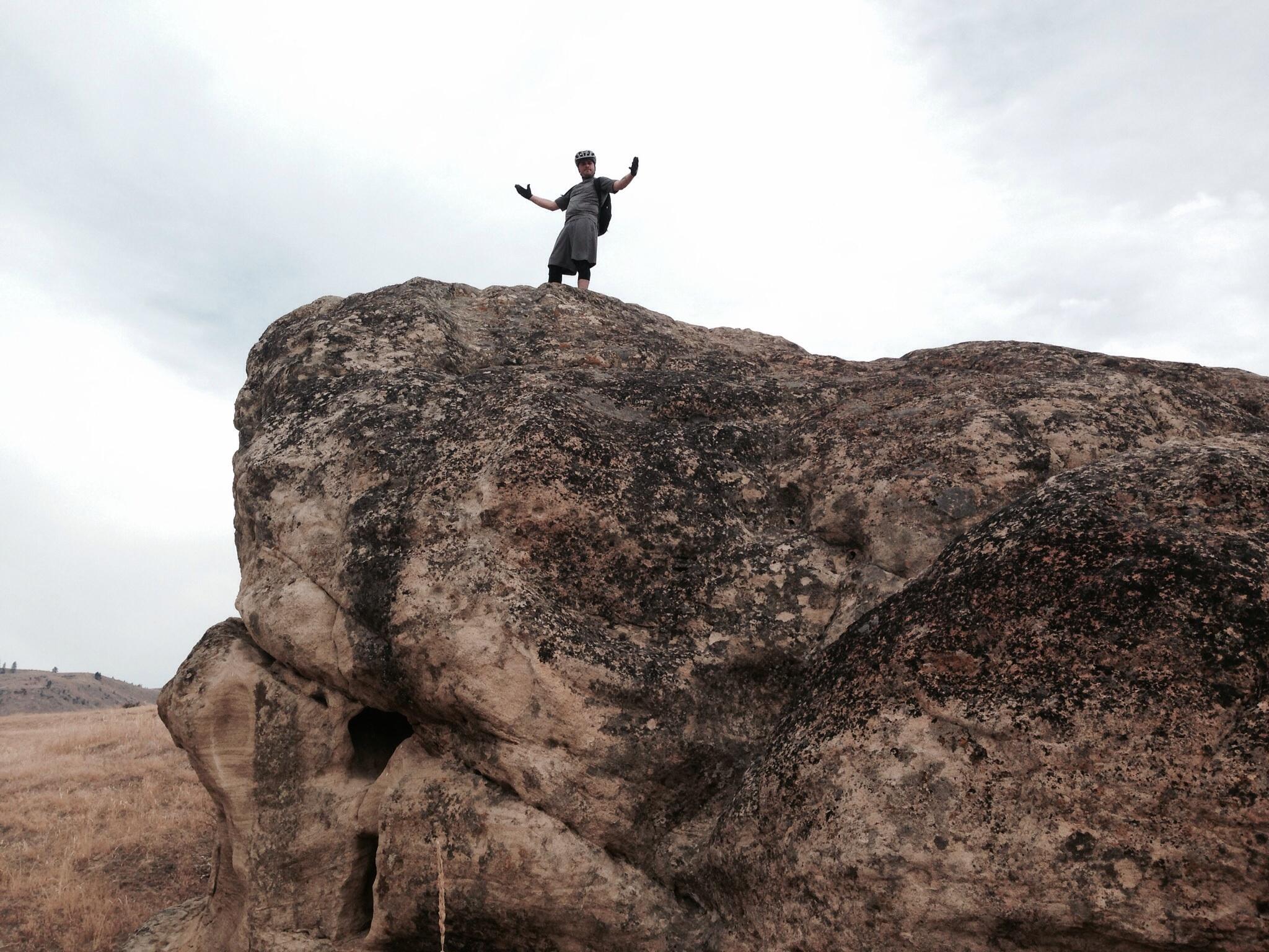 A person standing with outstretched arms on top of a large rock, set against a cloudy sky and dry landscape. The individual is wearing a helmet and casual clothing, exuding a sense of triumph or celebration. Sage Hills mountain bike trail.