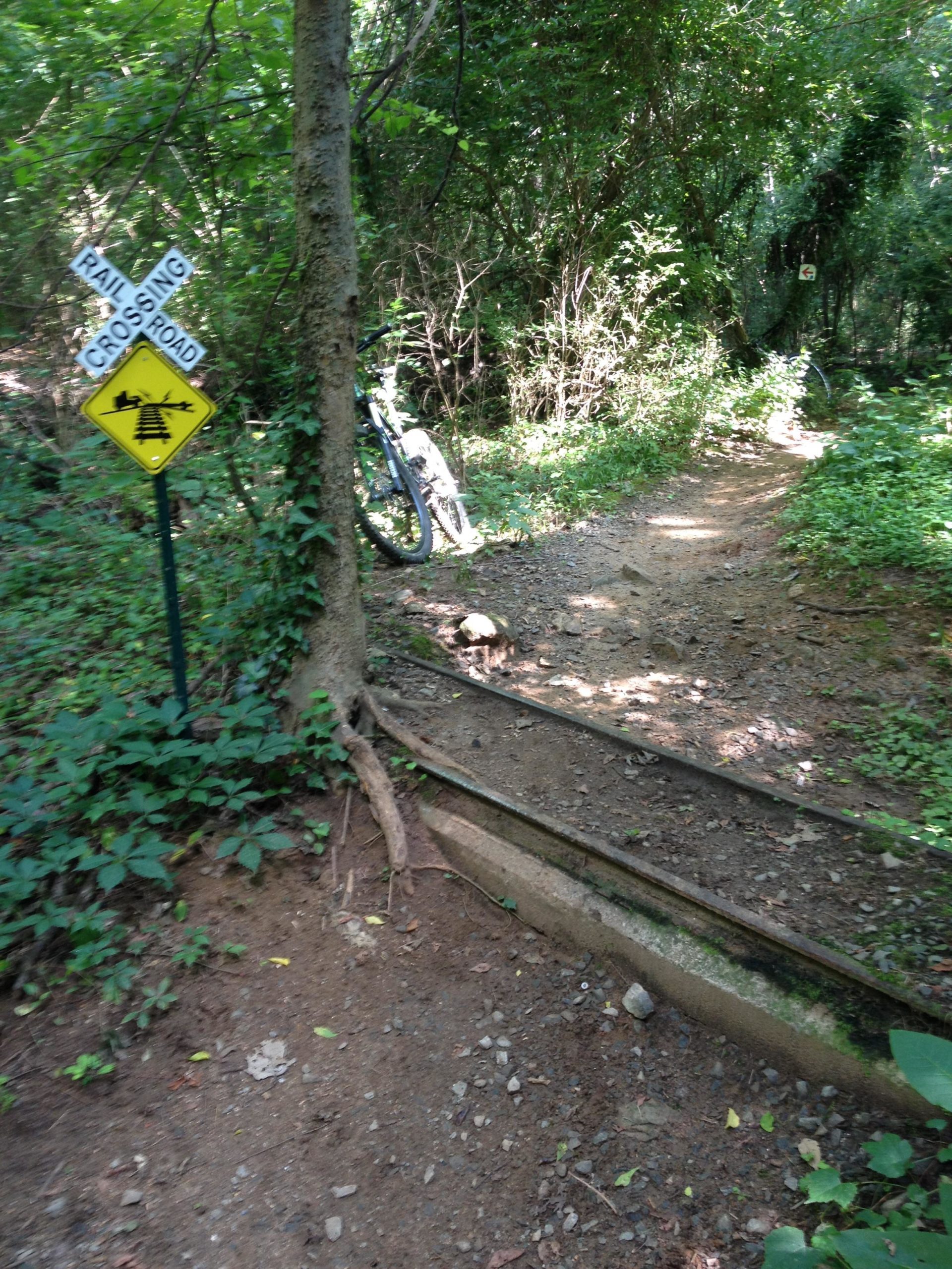 A narrow dirt path through a lush, green forest, featuring a bicycle leaning against a tree. A yellow sign warns of a railroad crossing, while another sign points to a road intersection. The surroundings are dense with trees and undergrowth, creating a natural and secluded atmosphere. Back Yard Trails mountain bike trail.