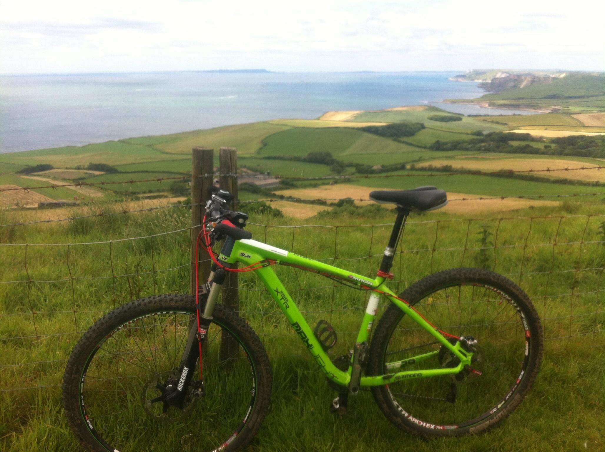 Kinesis FF29: A green mountain bike leaning against a wooden post, overlooking a scenic landscape with rolling green fields and a coastline in the background. The sky is partly cloudy, and the ocean is visible in the distance.