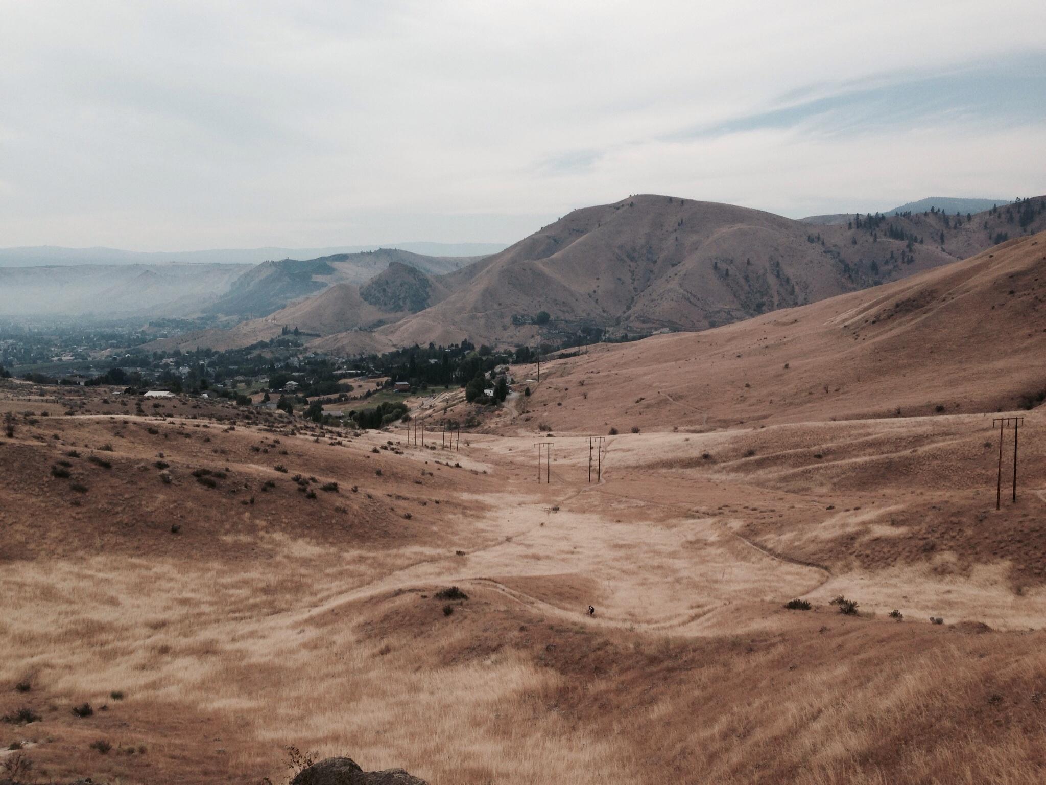 A panoramic view of a dry, hilly landscape under a cloudy sky, featuring rolling hills and sparse vegetation. In the foreground, tall grasses are visible, while the background shows a valley with scattered houses and power lines stretching across the terrain. The overall mood is tranquil, with a muted color palette of browns and greens. Sage Hills mountain bike trail.