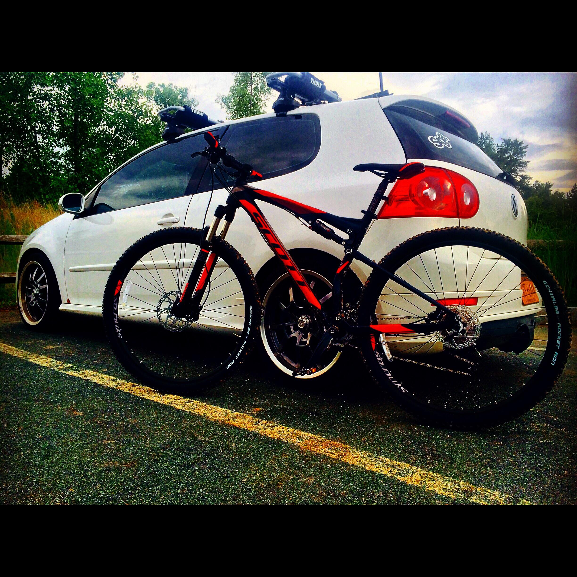 Scott Spark 960: A black and orange mountain bike is parked next to a white car in a scenic outdoor setting. The car has a bicycle rack on the roof and is positioned on a slightly faded parking lot line. Green trees and a partially cloudy sky are visible in the background.