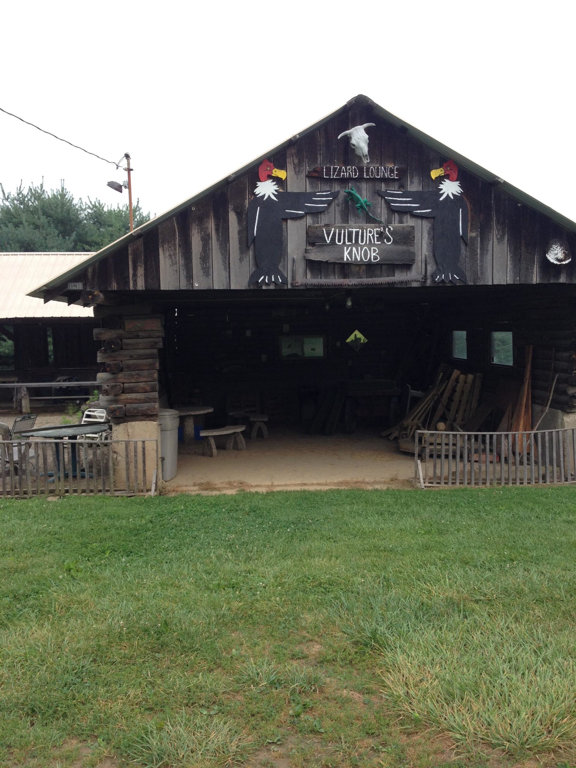 A rustic wooden structure labeled "Lizard Lounge" and "Vulture's Knob" features colorful painted decorations of birds, including a vulture, on the front. The building has an open area underneath, showcasing sandy ground and some furniture. Surrounding the structure is a grassy area, with hints of greenery and trees in the background. Vultures Knob mountain bike trail.