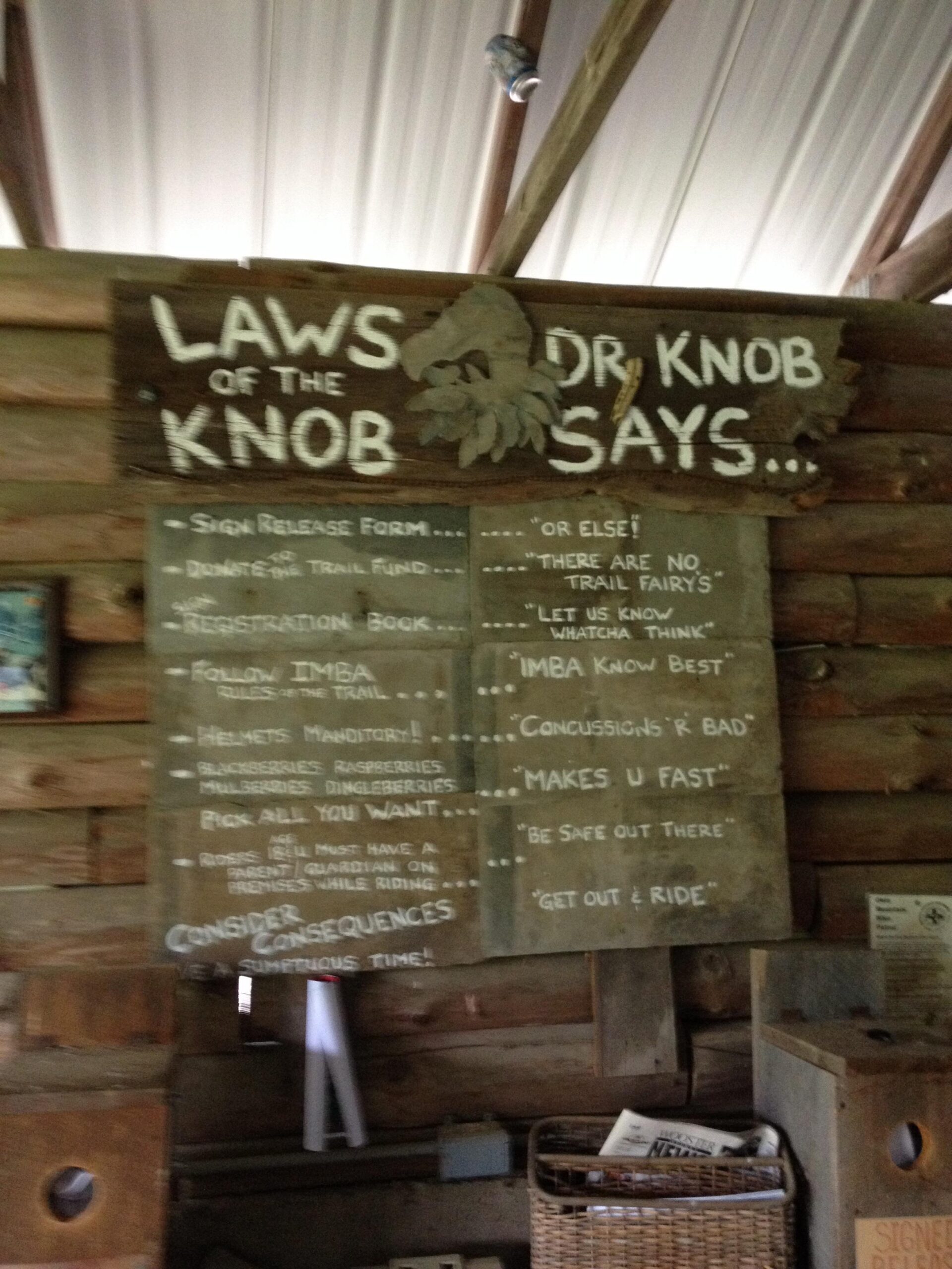 A wooden sign with the title "Laws of the Knob Dr. Knob Says" displayed prominently at the top. Below the title are various rules and guidelines for trail users, written in a casual style. The listed points include instructions about signing a release form, donating to the trail fund, helmet requirements, and reminders about the potential for concussions. Tips like "pick all you want" regarding berries, and playful remarks emphasizing safety and enjoyment are also included. The background features a rustic wooden wall and some additional items in the environment.  Vultures Knob mountain bike trail.