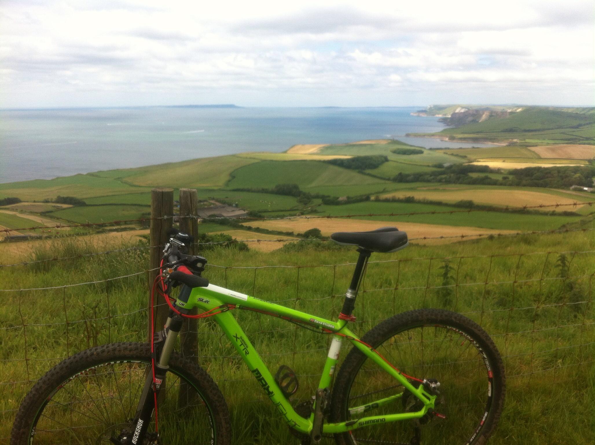 Kinesis FF29: A bright green mountain bike is leaning against a wooden fence, overlooking a scenic landscape of green fields and a shimmering blue sea under a partly cloudy sky. The coastline and distant hills are visible in the background, creating a stunning vista.