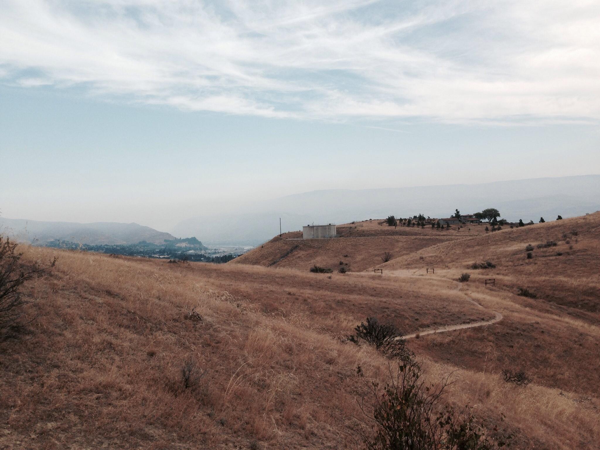 A panoramic view of a dry, hilly landscape under a cloudy sky. The foreground features golden grass and sparse vegetation, while a winding dirt path leads through the hills. In the distance, a small settlement is visible on a ridge, surrounded by more hills and a hazy horizon. The scene conveys a serene, open natural environment. Sage Hills mountain bike trail.