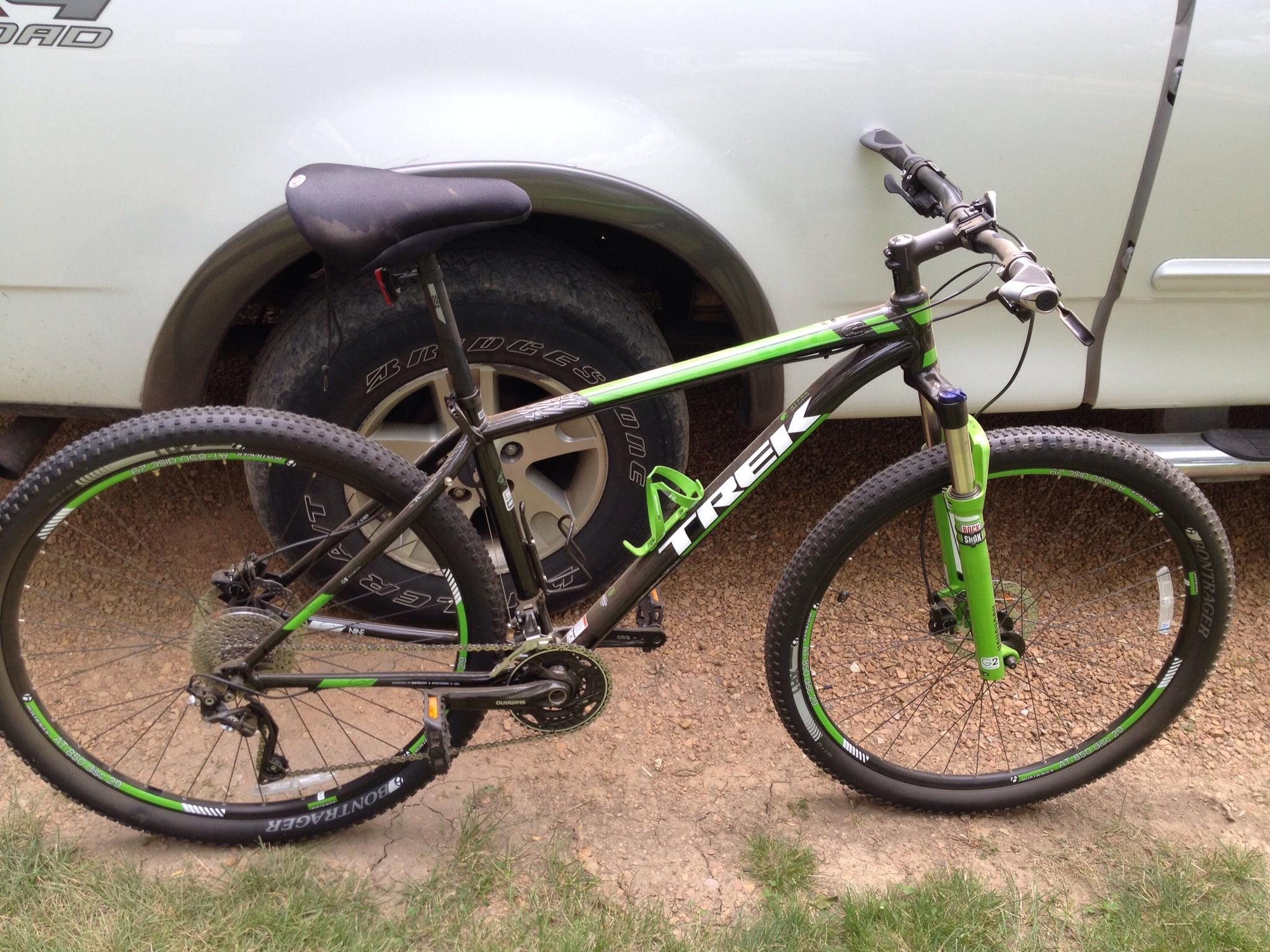 Trek X-Caliber 9: A green and black Trek mountain bike parked next to a vehicle, featuring knobby tires, a black seat, and a visible gear system. The background includes a gravel surface and some grass.