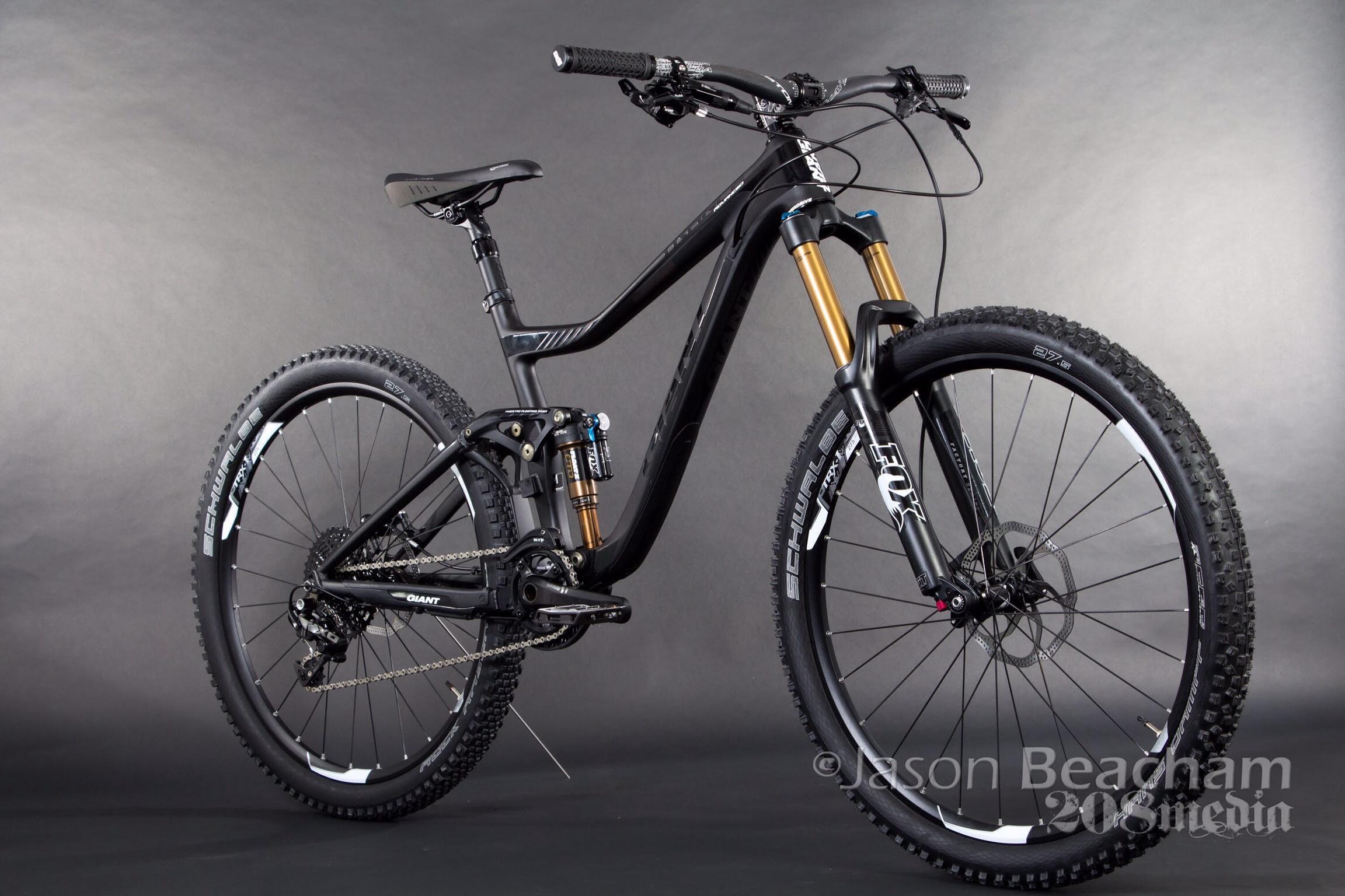 Giant Trance Advanced SX 27.5: A black mountain bike with gold suspension forks, featuring a sleek frame and wide tires. The bike is positioned against a gray background, showcasing its modern design and components.