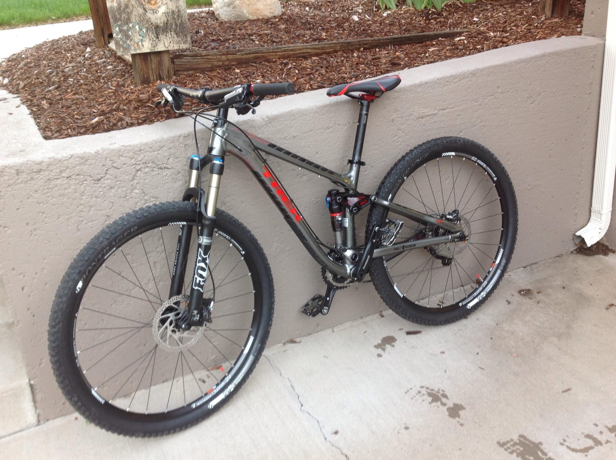 Trek Fuel EX 8: A black and red mountain bike leaning against a gray wall, featuring front suspension forks and wide tires. The bike has disc brakes, and the surrounding pavement shows light footprints. In the background, there is a wooden structure and mulch landscaping.