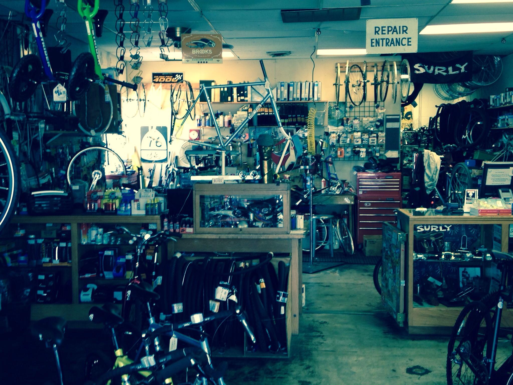 A bustling bike repair shop filled with various bicycles, tools, and accessories. The interior features wooden counters displaying bike components and repair tools, with an assortment of hanging bicycles and wheels on display. Brightly colored signage indicates the repair entrance, and the shop is warmly lit, creating a welcoming atmosphere for cyclists.