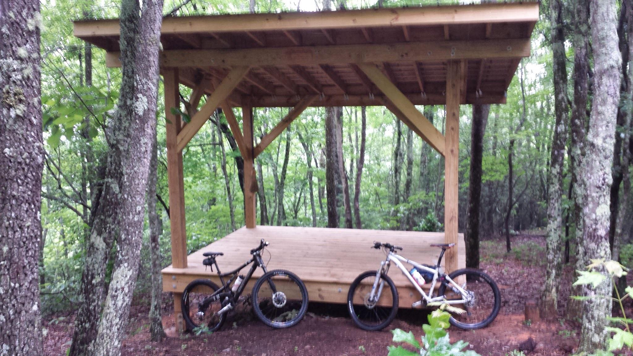A wooden bike shelter with a spacious platform is surrounded by lush green trees in a forest. Two mountain bikes, one black and one white, are leaning against the shelter, indicating a perfect spot for cyclists to rest and enjoy nature. Bracken Preserve mountain bike trail.
