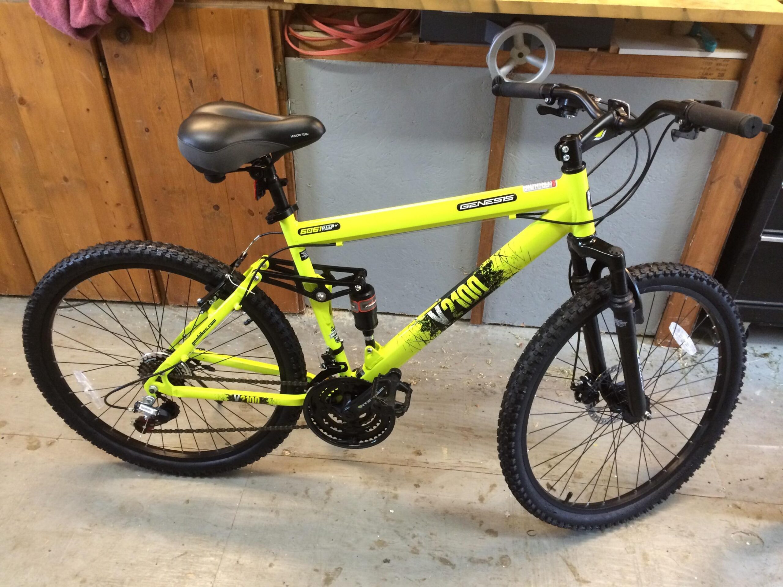 Genesis V2100: A bright yellow mountain bike with black tires and handlebars, featuring a black seat. The bike is positioned in a garage setting with wooden walls and a workbench in the background.