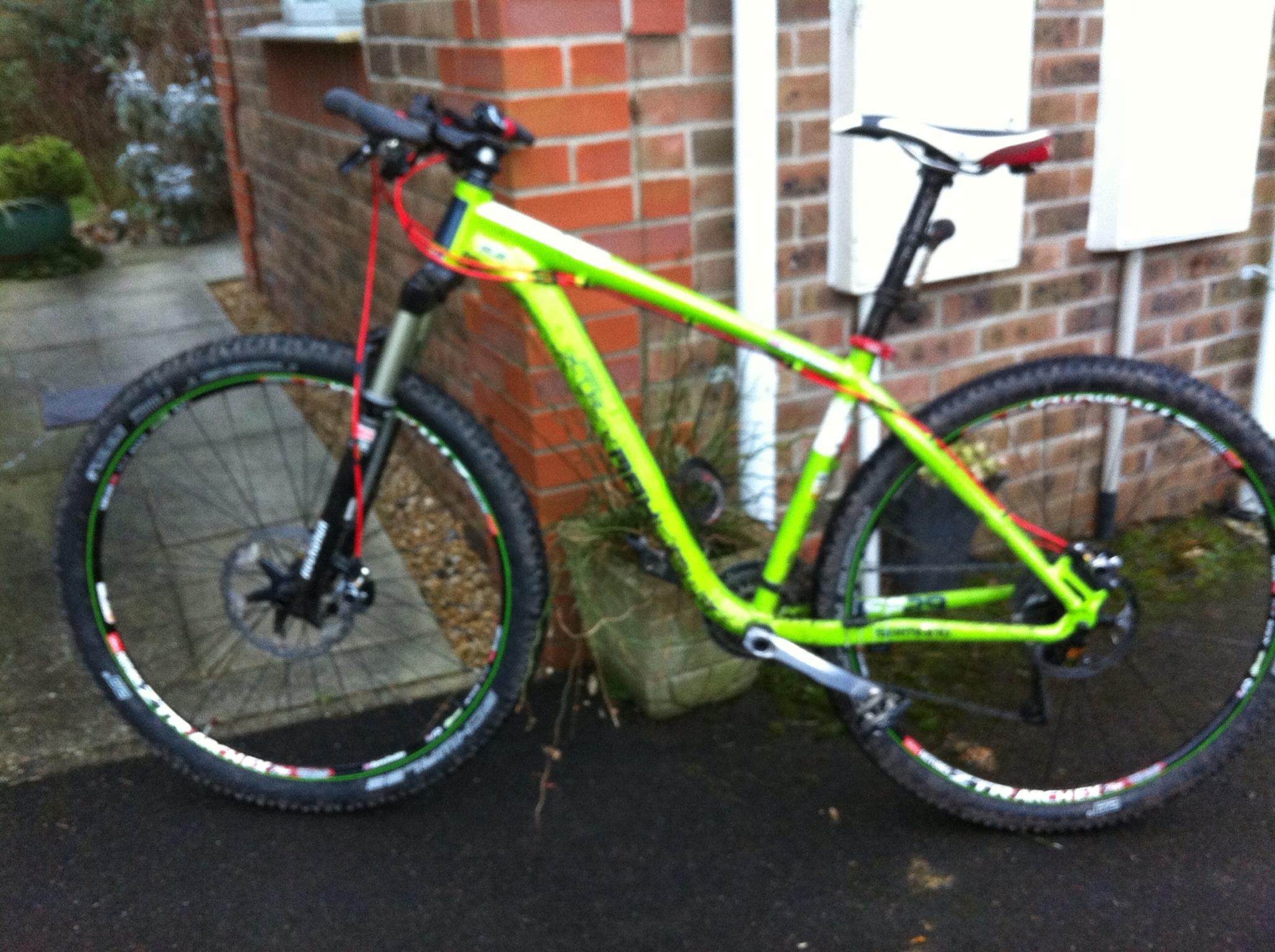 Kinesis FF29: A brightly colored green mountain bike parked next to a brick wall, with a concrete path and a potted plant in the background. The bike features large, knobby tires, disc brakes, and a prominent front suspension. The handlebars are equipped with controls, and the seat is positioned high on the frame.