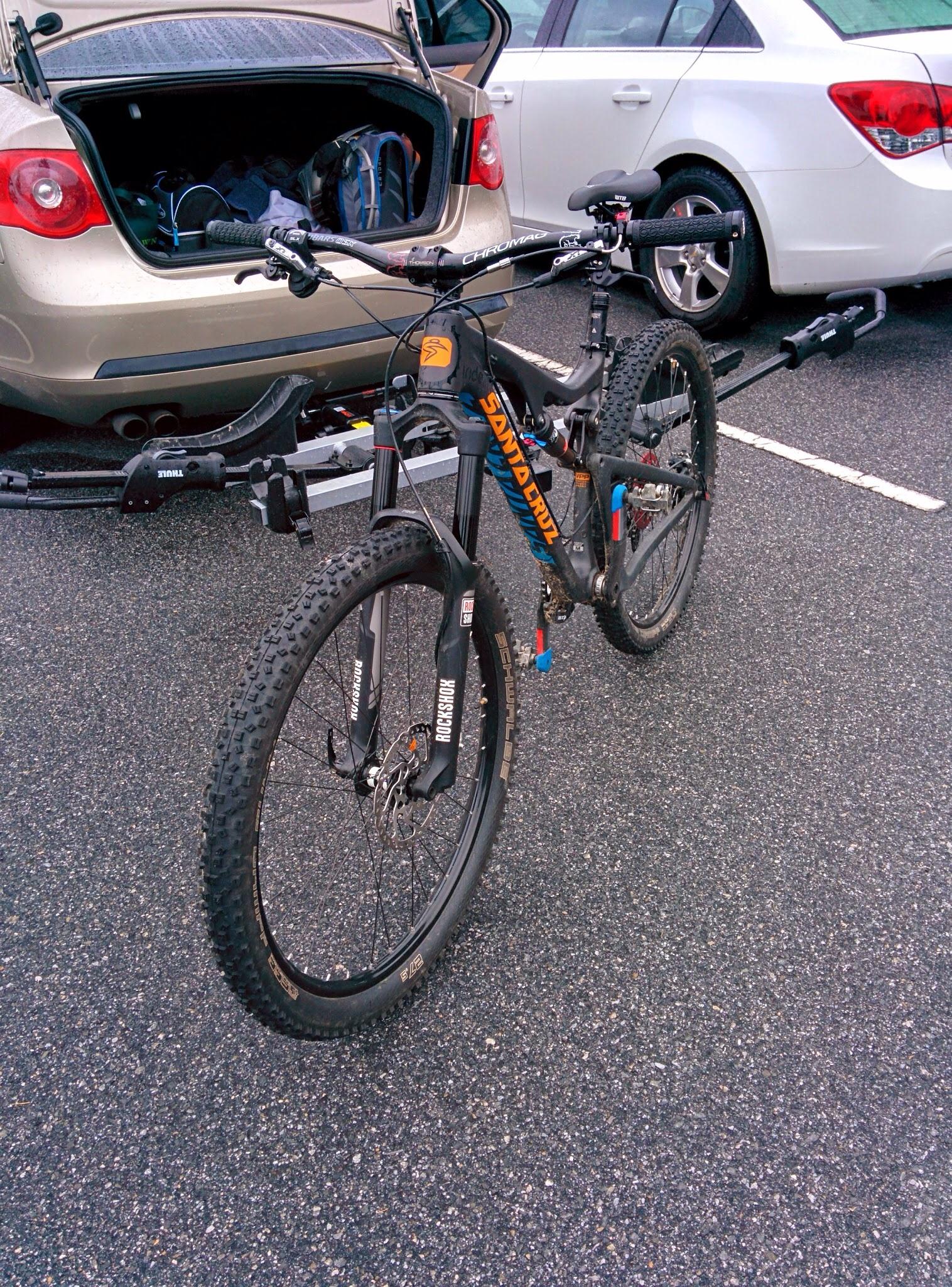 Santa Cruz Bronson: A mountain bike with a prominent blue and orange "Santa Cruz" logo parked in front of a car's open trunk, revealing bags and equipment inside. The bike is mounted on a hitch rack, and the scene is set in a parking lot with asphalt pavement.