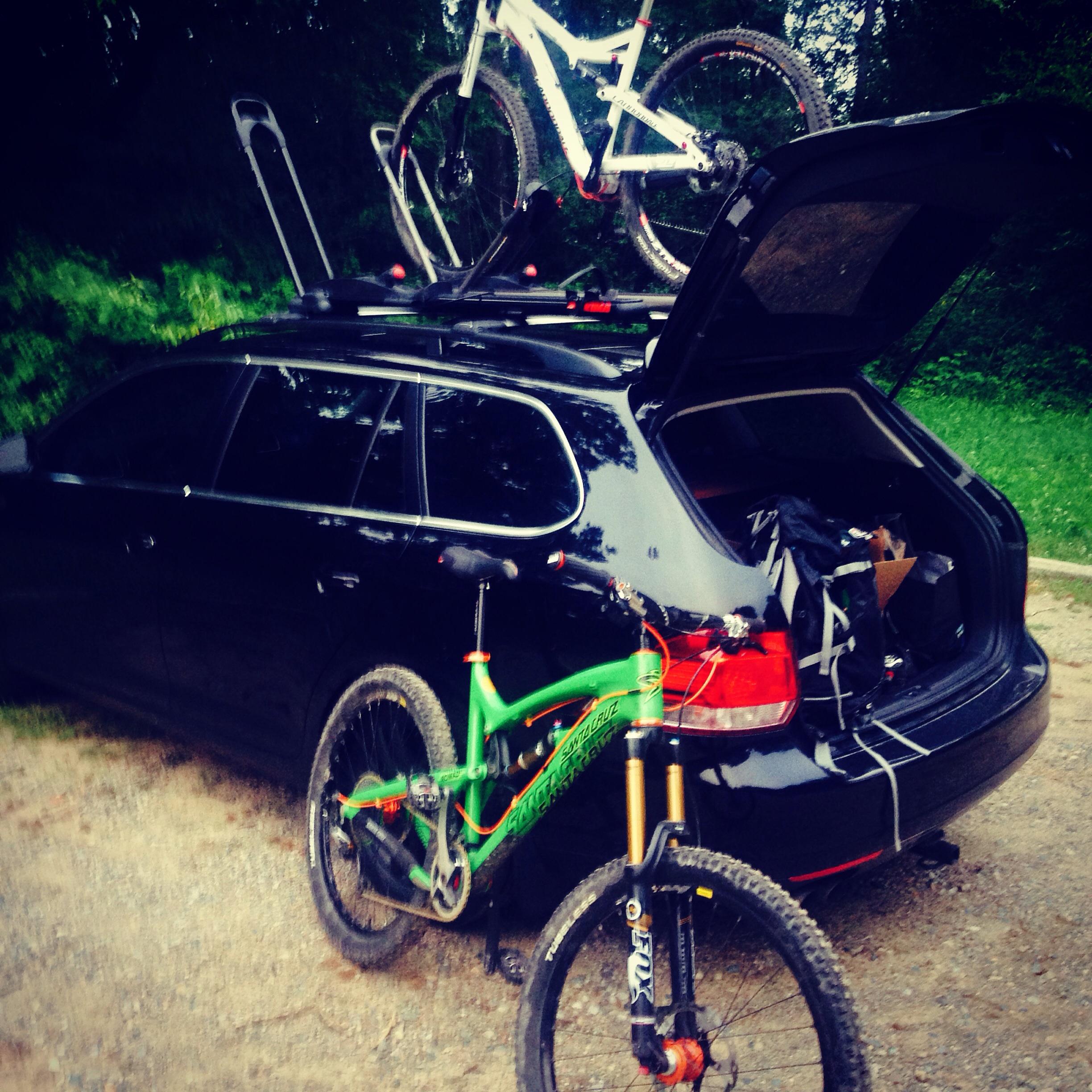 Santa Cruz Nomad Carbon: A black SUV with its trunk open, showing various items inside. On the roof, a white mountain bike is secured, while a vibrant green mountain bike is positioned next to the vehicle on the ground. Surrounding greenery suggests an outdoor setting, possibly near a trail or park.
