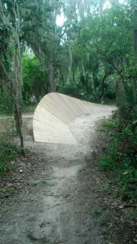 A wooden ramp or bridge curving over a dirt path in a lush, green forested area. Tall trees and hanging moss surround the pathway, creating a natural, serene environment. The ramp appears to blend into the landscape, leading the viewer's eye further along the trail. Loyce E. Harpe Park mountain bike trail.