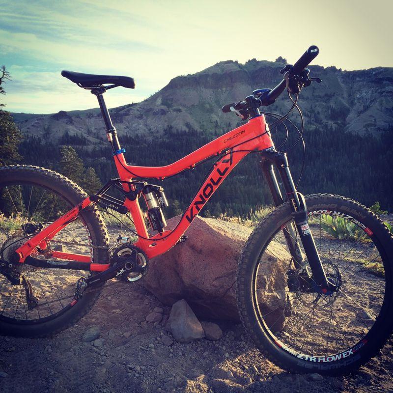 A vibrant orange mountain bike is positioned next to a large rock on a dirt trail, with a backdrop of lush green trees and rocky hills under a clear sky. The bike features a sturdy frame and thick tires, indicating it is designed for off-road use. Hole In The Ground mountain bike trail.