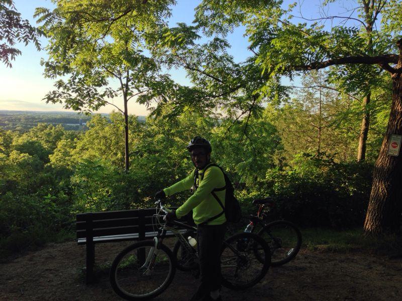 A cyclist in a bright yellow jacket stands next to a mountain bike in a wooded area, with a scenic view of lush green hills in the background. A bench is visible nearby, and the scene is illuminated by soft natural light. Emma Carlin mountain bike trail.