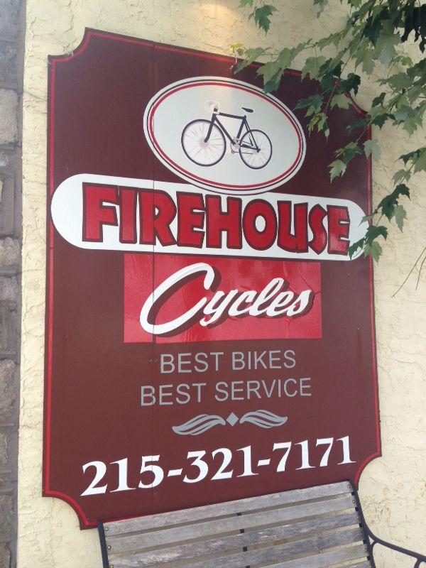 Sign for Firehouse Cycles featuring a bicycle graphic, with the text "Firehouse Cycles" prominently displayed. Below, it states "Best Bikes, Best Service" and includes a phone number: 215-321-7171. The sign is mounted on a light-colored wall with some leafy greenery nearby.