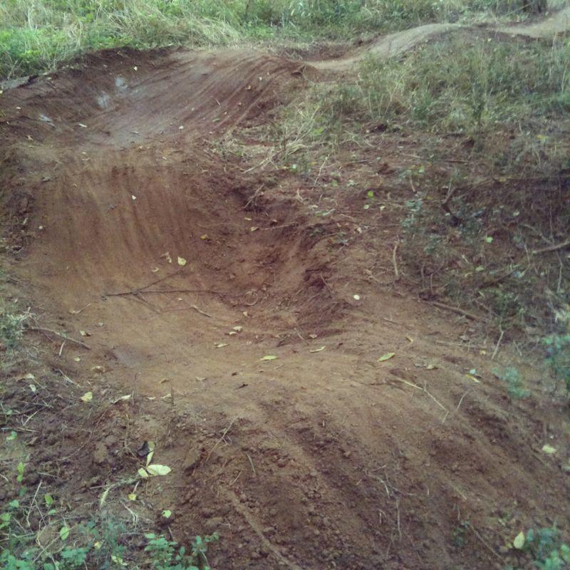 A dirt jump ramp located in a grassy area, showing a smooth, curved surface with a slight incline and a flat base. The surroundings are filled with tall grass and scattered leaves, indicating it