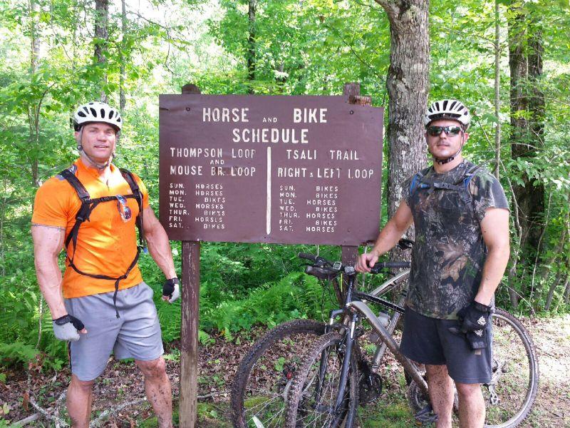Two men stand near a wooden sign that displays the horse and bike schedule for various trails, including Thompson Loop and Tsali Trail. One man is wearing an orange shirt and the other is in camouflage, both equipped with helmets and bikes. The background features a lush green forest. Research and Technology Park (ERC) mountain bike trail.